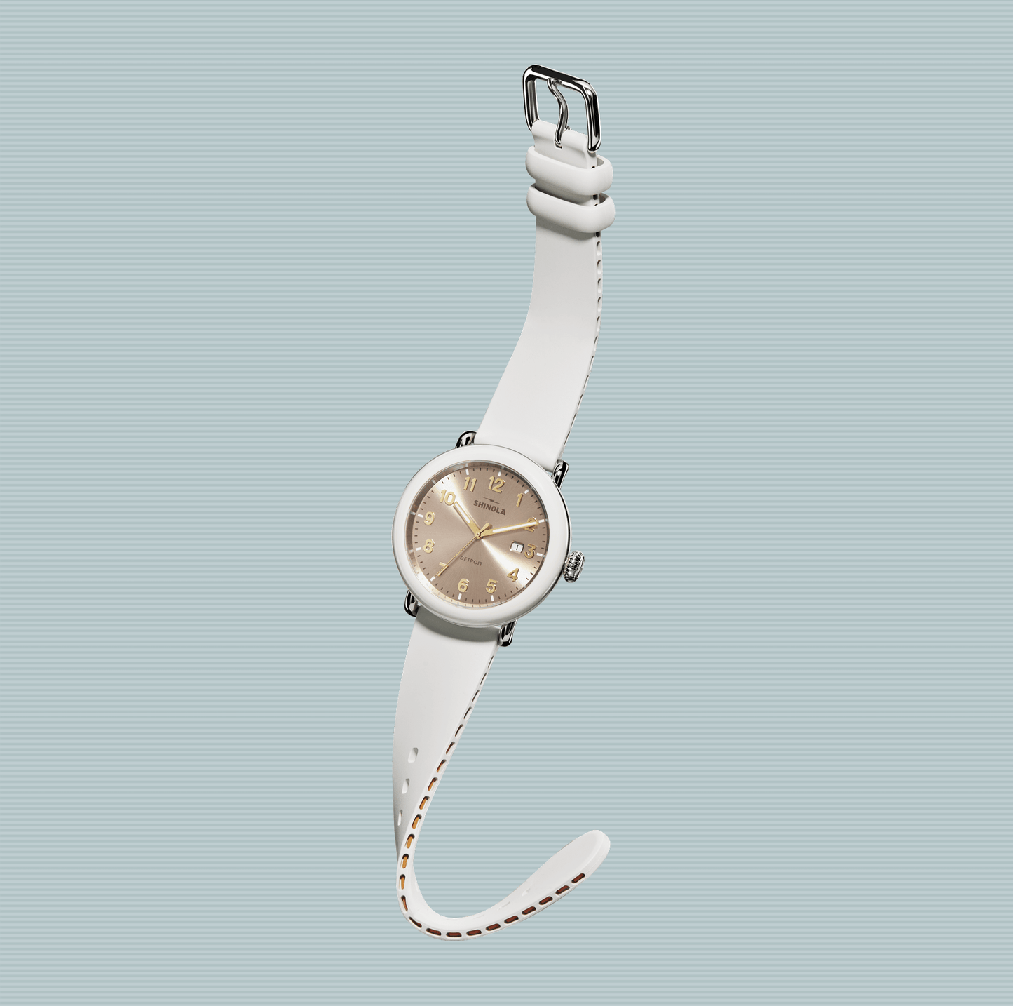 The Shinola Detrola Watch - Nude, with a beige dial and gold numbers on a white strap, is shown against a light blue striped background, highlighting its classic American design.
