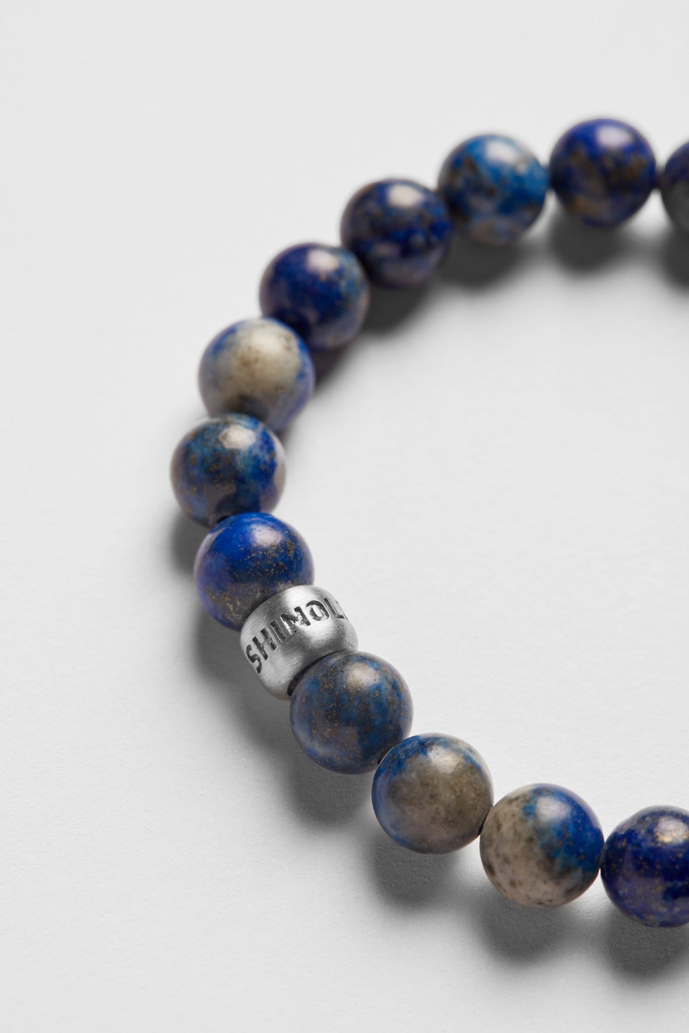 A close-up of the Shinola 8mm Beaded Lapis Bracelet, featuring round blue lapis beads and a single silver COURAGE-engraved bead, displayed on a light gray background.
