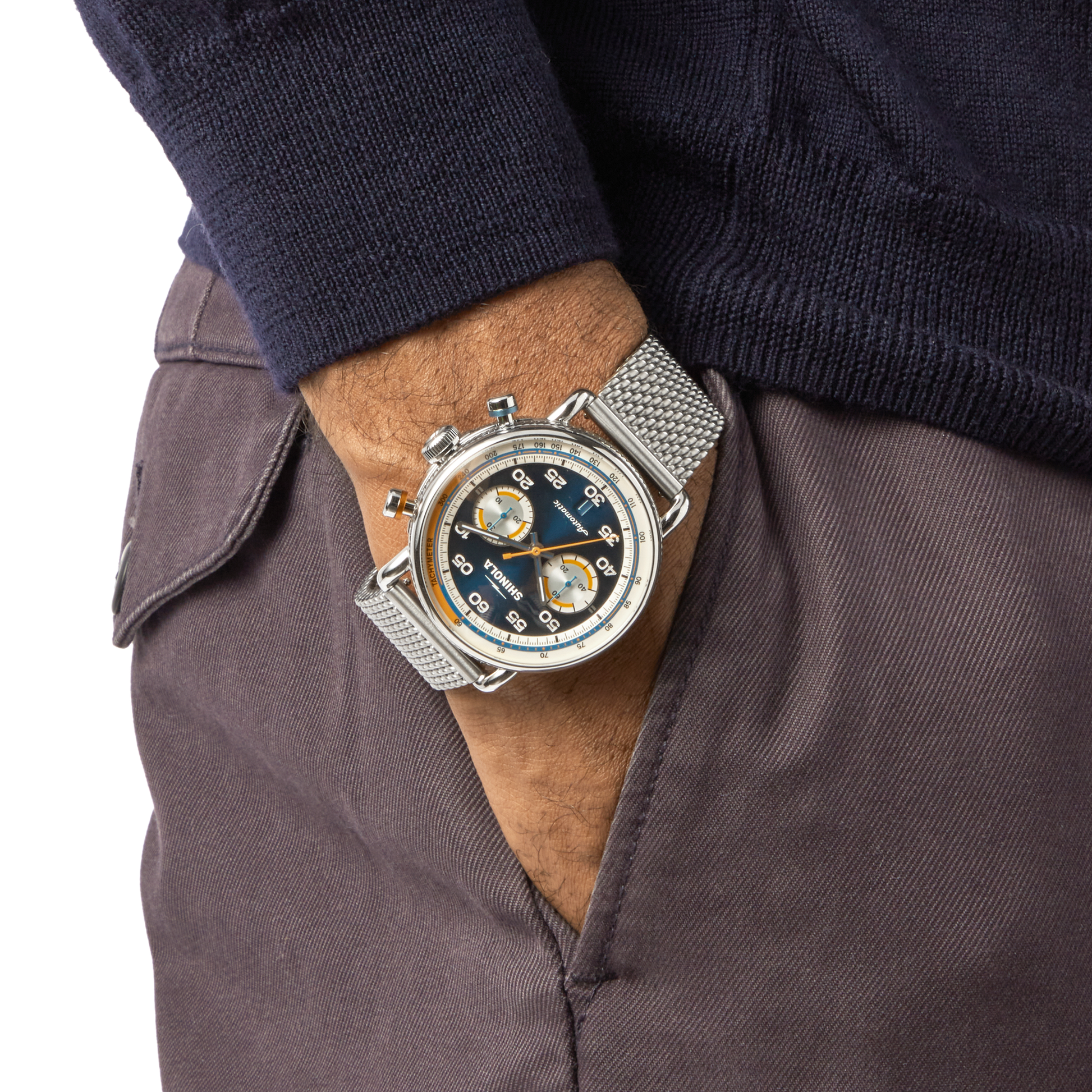 A person in a dark sweater and brown pants has their hand in their pocket, displaying the Shinola Canfield Speedway Open Road Automatic Chronograph Watch - Blue with a silver metal mesh strap on their left wrist.