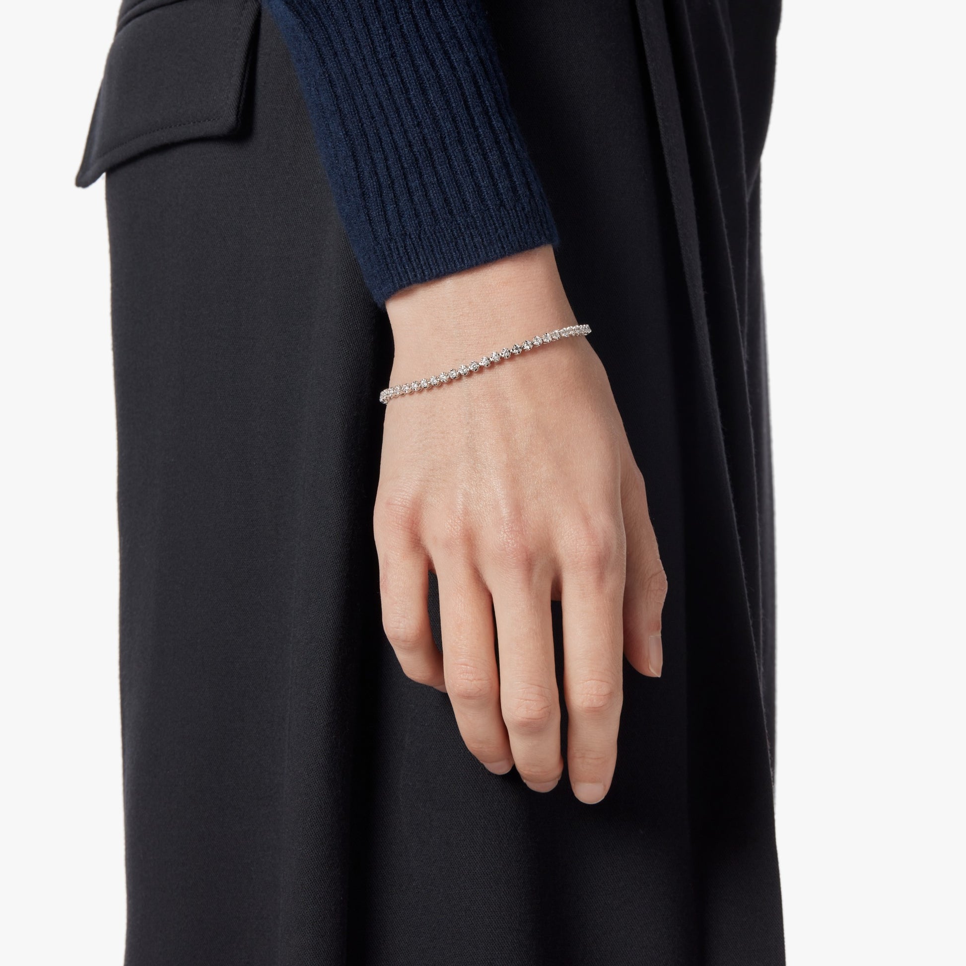 A person in a dark long-sleeve top and skirt has their left arm by their side, elegantly displaying the Shinola 2 Carat 7" Diamond Tennis Bracelet in White Gold on their wrist.