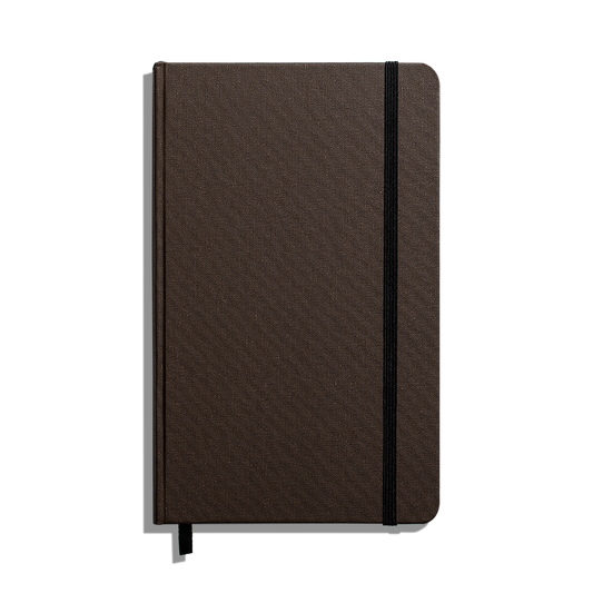The Shinola Medium Hard Ruled Journal (5.25" x 8.25") is a closed brown hardcover notebook with rounded corners, a black elastic band, acid-free paper, and a black ribbon bookmark, displayed on a white background.