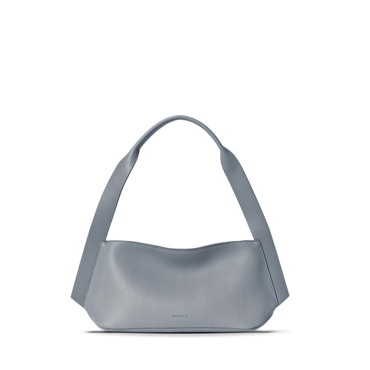 The Shinola Small Derby Shoulder Bag - Mist is a minimalist light blue petite shoulder bag crafted from natural leather, with a wide, curved strap, smooth structured body, and subtle embossed logo near the bottom center. Displayed on a white background.
