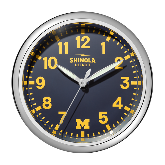 A 14" Shinola Runwell Clock with a silver frame, blue face, yellow numbers and hands. The clock reads 1:09:35, shows Shinola Detroit at the top, and has a yellow M for the University of Michigan at the bottom.