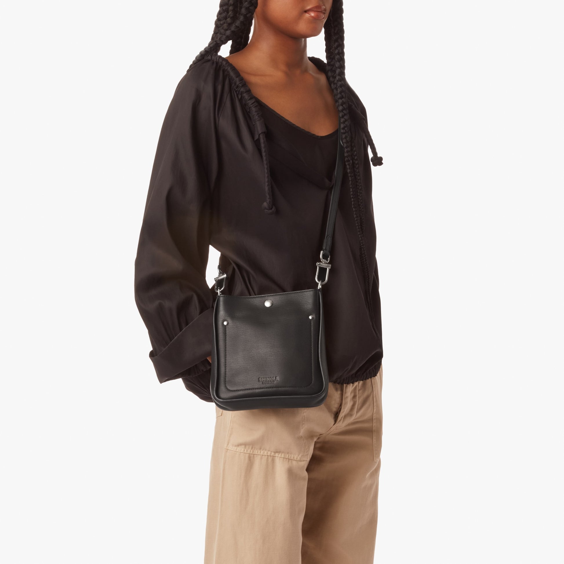 A woman in a loose black blouse and beige pants stands against a white background, wearing the Shinola Mini Pocket Crossbody Bag - Black with an adjustable leather strap over her shoulder.