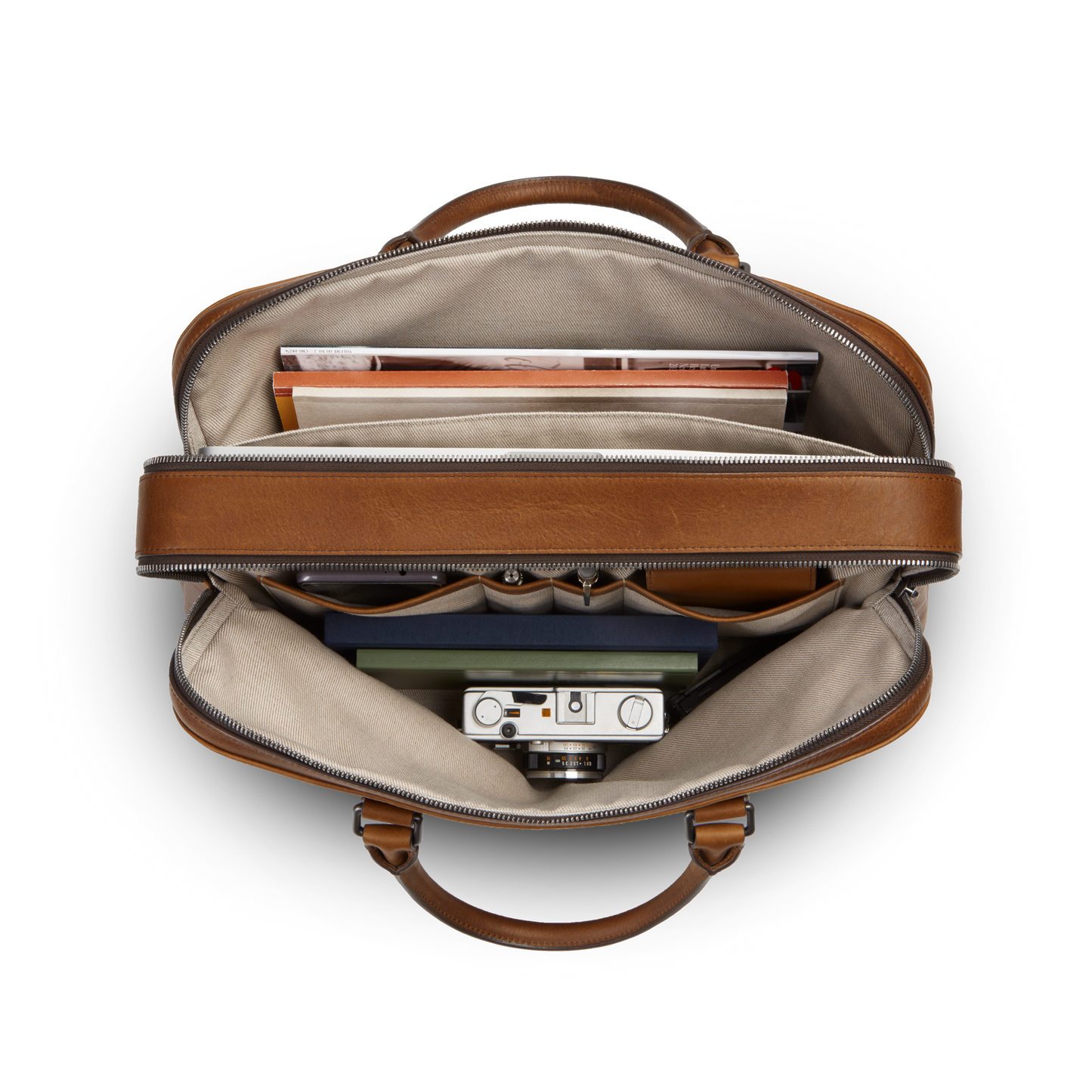 Top view of an open Shinola Double Zip Briefcase - Medium Brown shows books, a folder, a camera, and small items neatly organized inside beige fabric compartments.