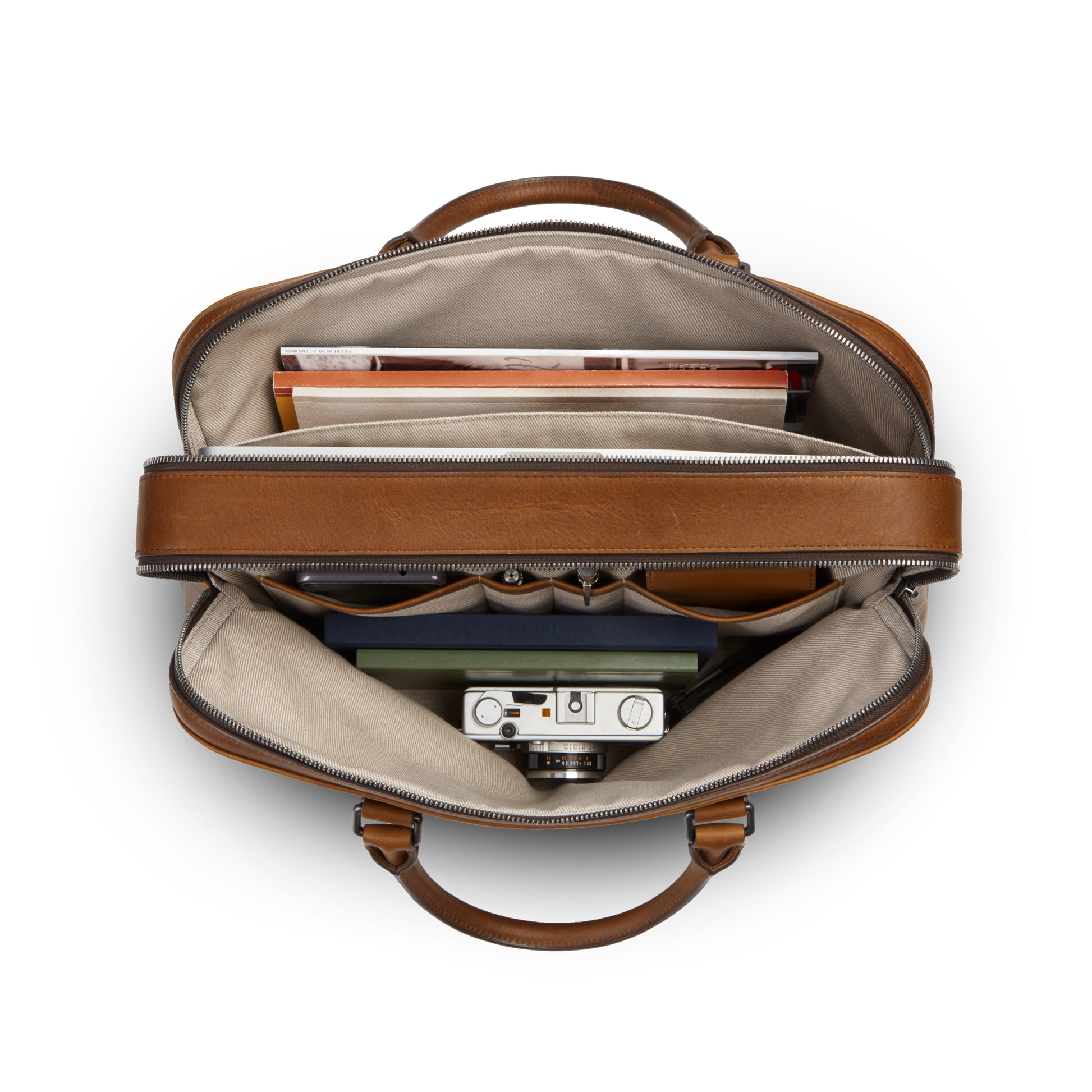 Top view of an open Shinola Double Zip Briefcase - Medium Brown shows books, a folder, a camera, and small items neatly organized inside beige fabric compartments.