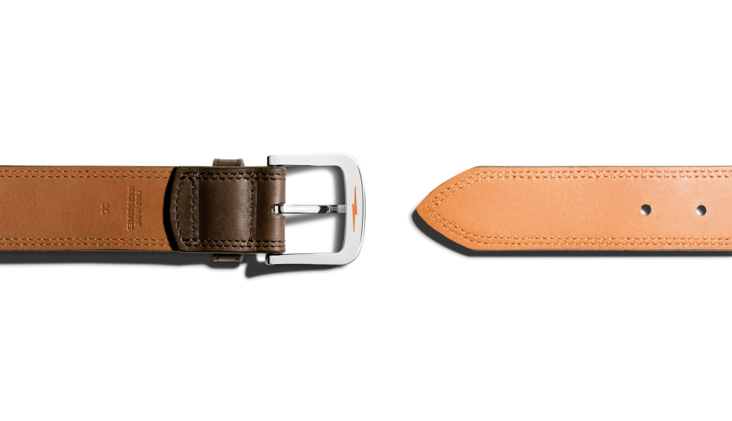 The Shinola 1 1/2" Double Stitch Belt in Deep Brown lies horizontally on a white background, displaying its silver buckle and brown strap to the left, with the tan tip and adjustment holes on the right.