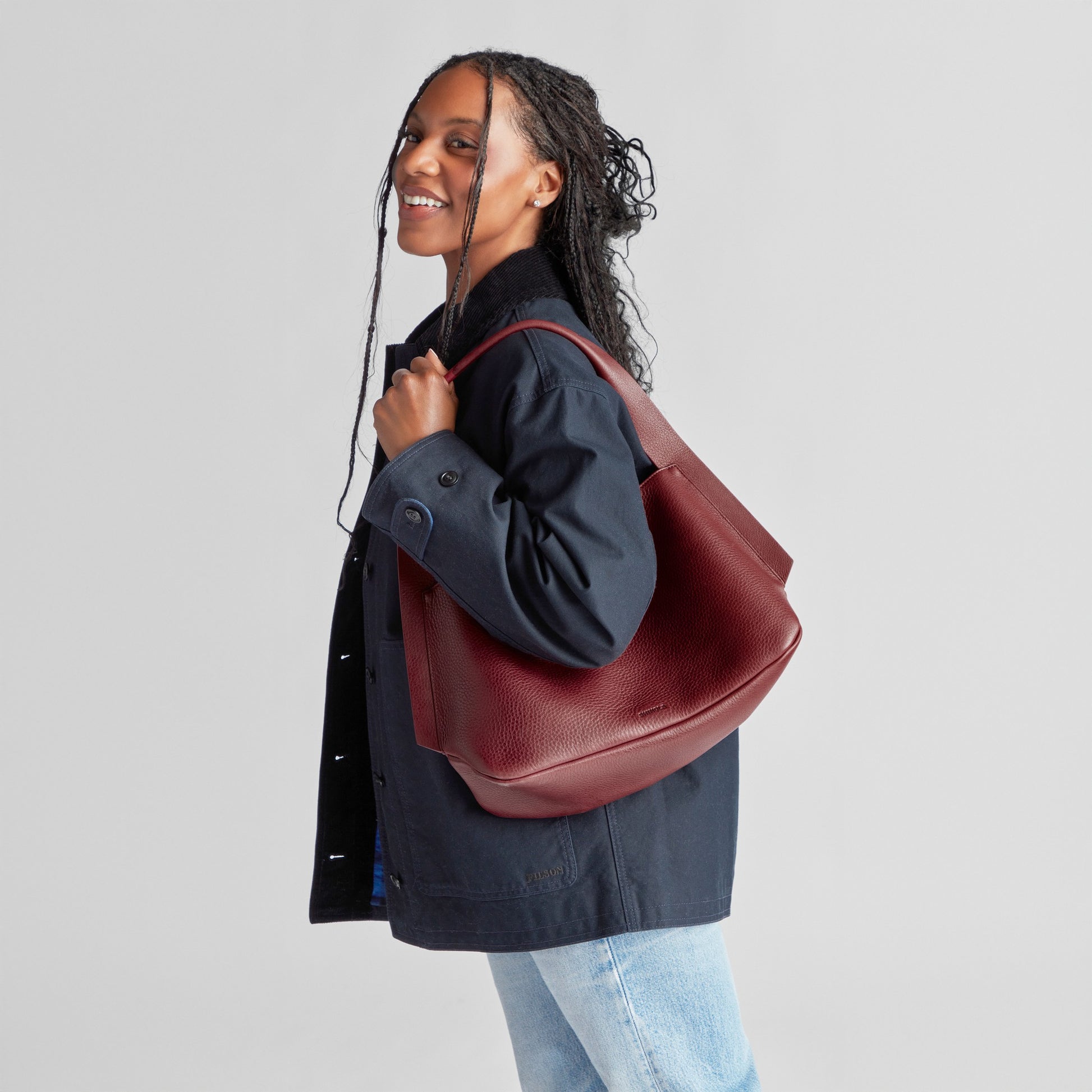 A smiling woman with long braids wears a navy jacket and light blue jeans, holding a Shinola Medium Derby Shoulder Bag in Ruby over her left shoulder against a plain light gray background.