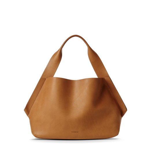 The Shinola Derby Shoulder Bag - Tan is a large tote with wide handles, a minimal unstructured silhouette, and a discreet leather card pocket, shown standing upright against a white background.
