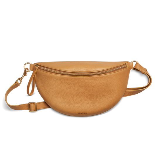 The Shinola Sling Bag - Honey, featuring a tan half-moon shape, zipper closure, adjustable leather strap, and gold-tone hardware, is shown against a white background.