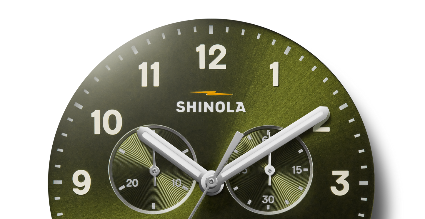 Close-up of the Shinola Canfield Chronograph Watch - Dark Olive, featuring a green dial with white hour markers, two subdials, three silver hands at 10:09:39, and the Shinola logo below 12 o'clock.