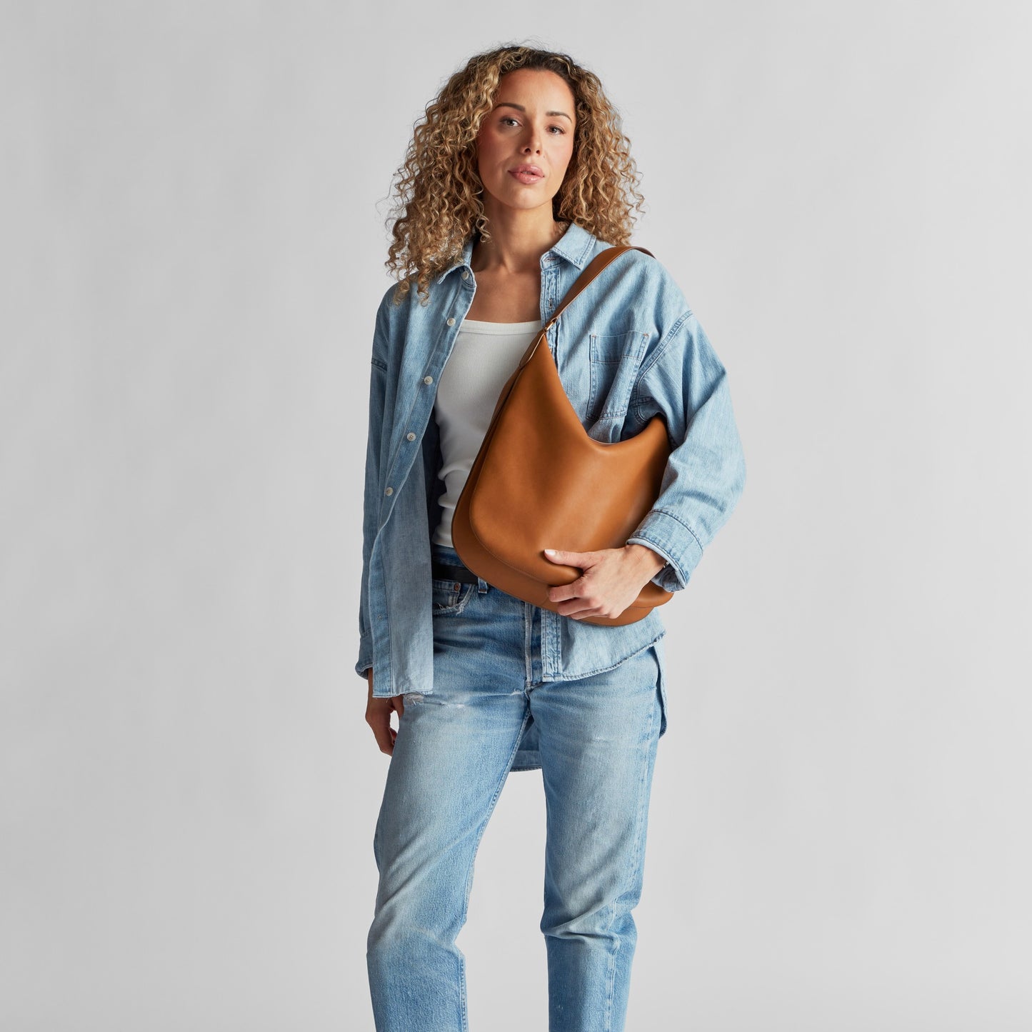 A person with curly hair wears a light blue denim jacket and jeans over a white top, holding the Shinola Bixby Shoulder Bag - Tan with accordion-style construction, standing against a plain light gray background.