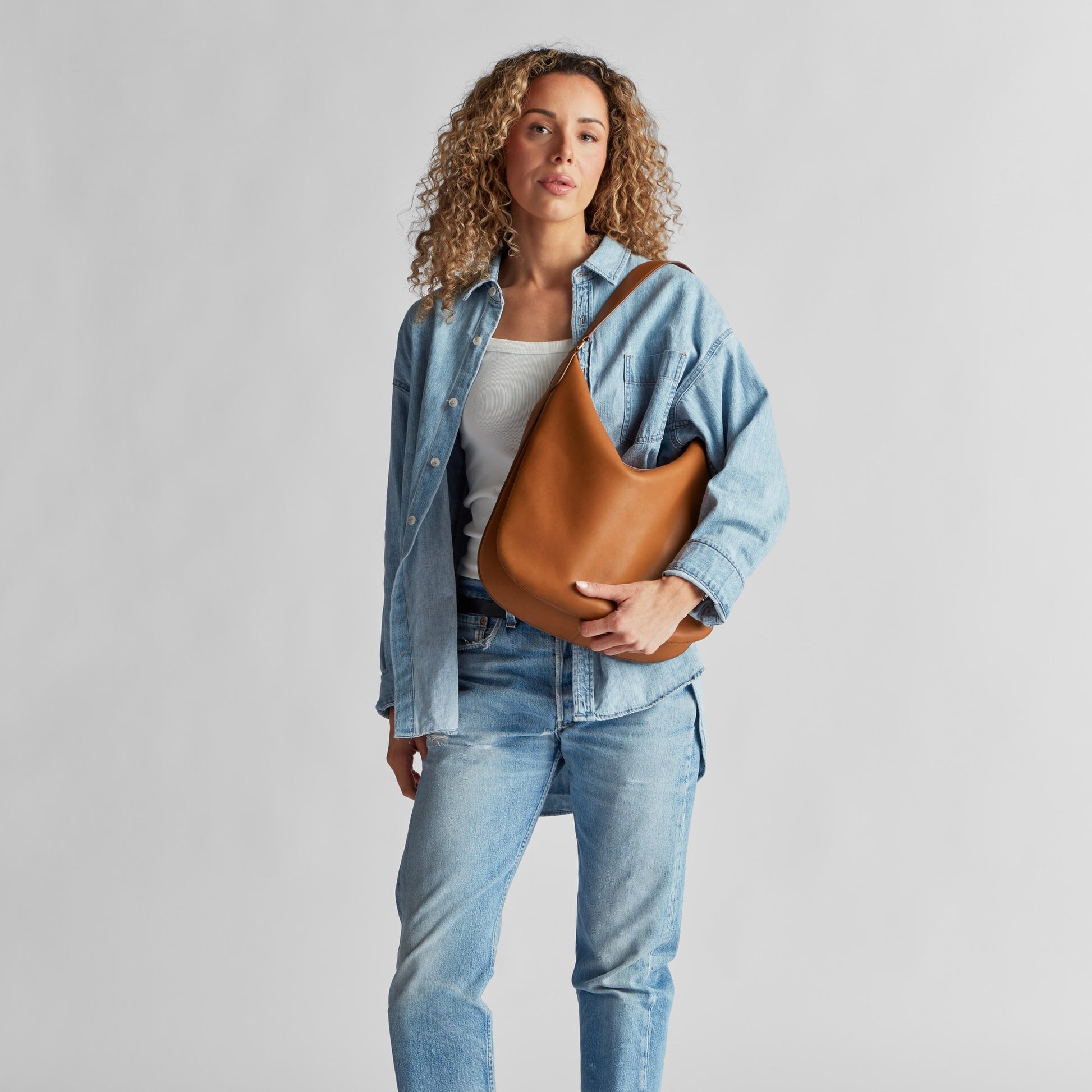 A person with curly hair wears a light blue denim jacket and jeans over a white top, holding the Shinola Bixby Shoulder Bag - Tan with accordion-style construction, standing against a plain light gray background.