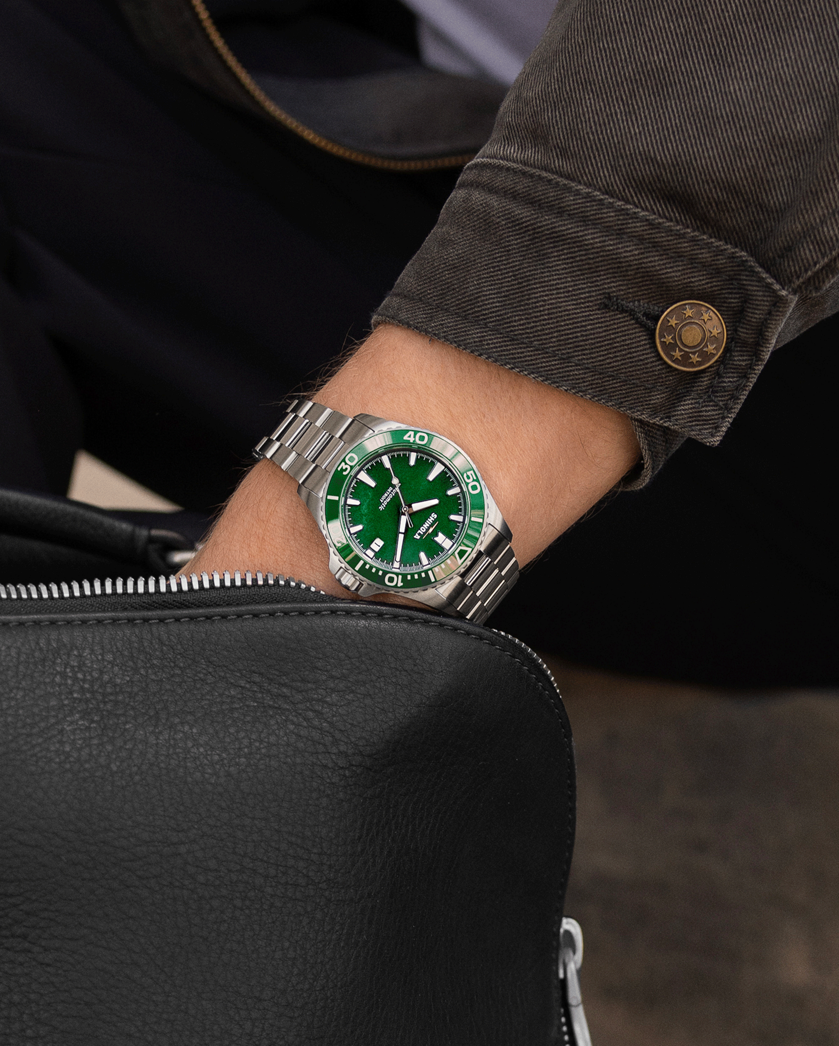 A person wearing a Shinola Lake Ontario Monster Automatic Dive Watch with a green face and bezel rests their hand on a black leather bag, dressed in dark pants and a brown jacket.