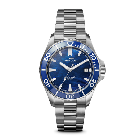 The Lake Michigan Monster Automatic Dive Watch by Shinola features a stainless steel case, blue and white dive bezel, blue dial, silver hour markers, date window at 3 o’clock, and showcases the Shinola brand on its automatic movement.