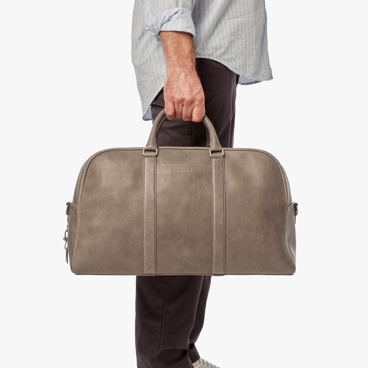 A person in a light blue shirt and dark pants holds the Shinola Runwell Duffle Bag - Stone by its handles against a plain white background.