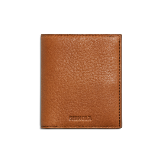 The Shinola Coin Bifold - Tan is a slim, textured natural grain leather wallet with SHINOLA embossed near the bottom center, shown closed on a plain white background. It offers refined style and lasting durability.