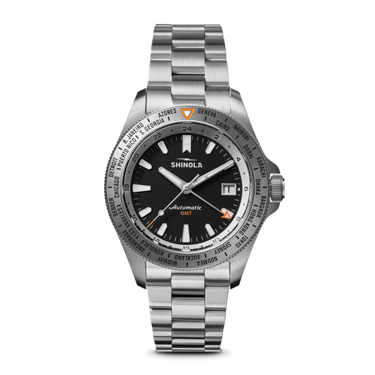 The Shinola Monster Worldtimer GMT Watch features a black dial, silver hour markers, date window at 3 o’clock, world timer bezel with cities, orange GMT hand, and a metal link band—perfect for travelers.