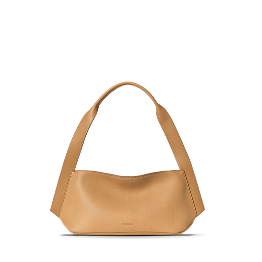 The Small Derby Shoulder Bag - Honey by Shinola features natural leather in a petite, curved silhouette with a wide strap and minimal design, subtly embossed with the Shinola logo near the bottom center against a white background.