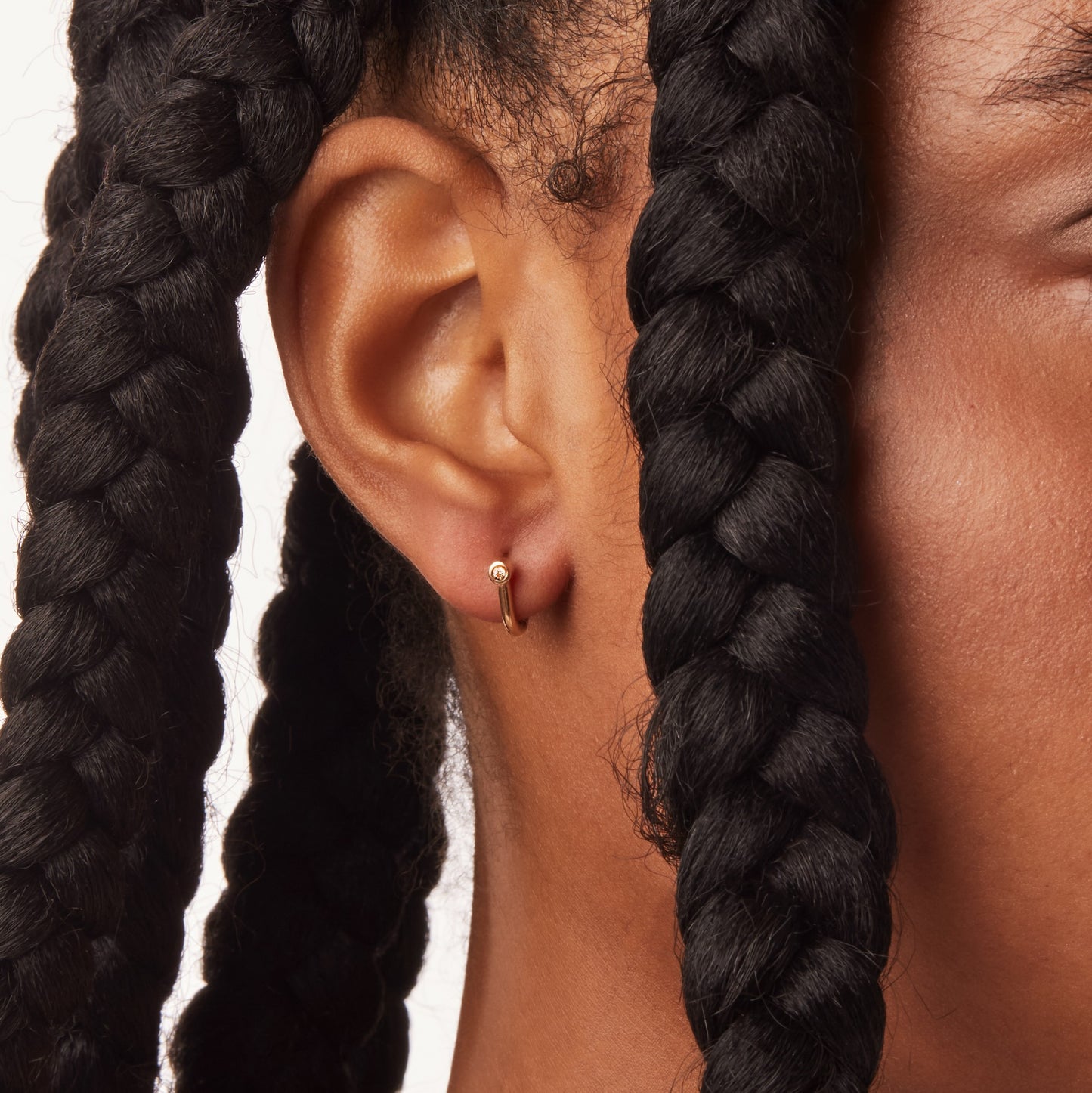 Close-up of a person with medium to dark skin and thick black braided hair wearing Shinola Diamond Bike Lock Huggie Earrings in yellow gold. The petite diamond earrings stand out against a white background.