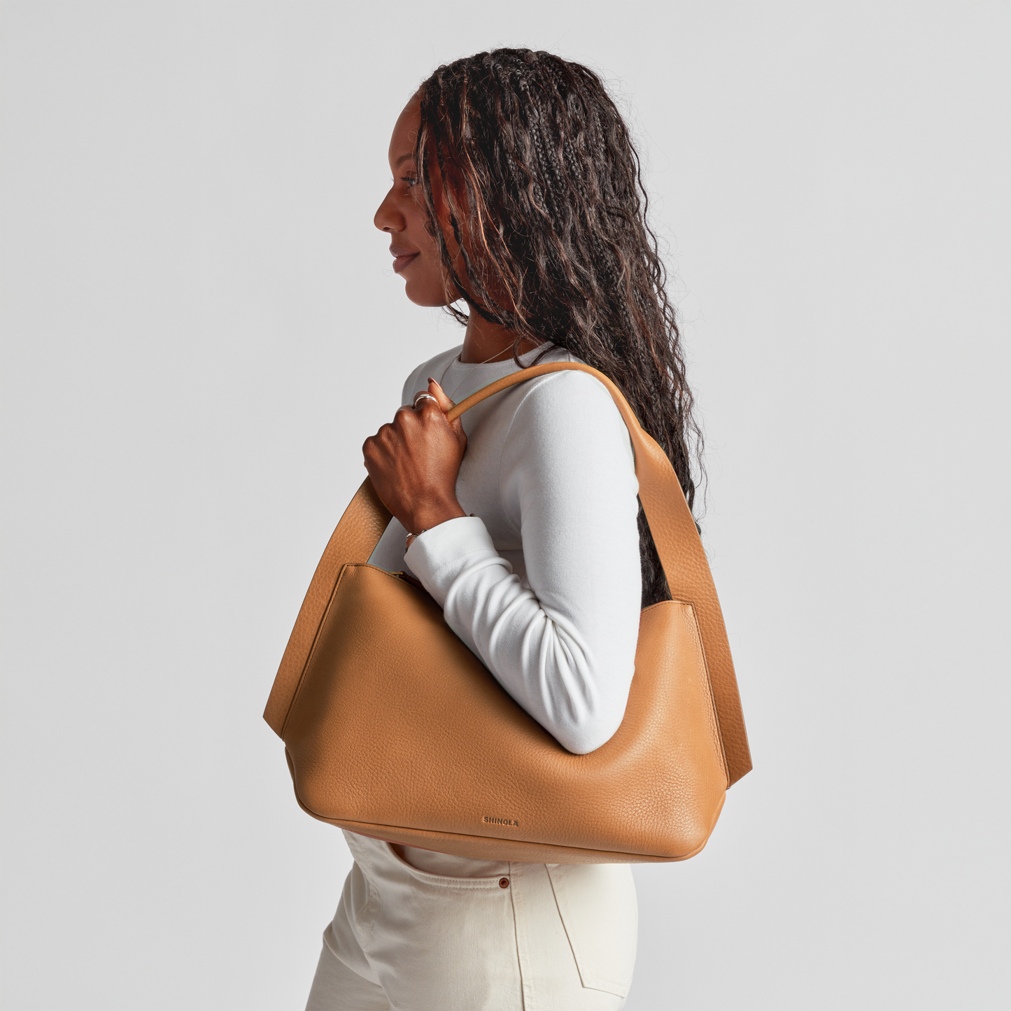 A woman with long curly hair, wearing a white long-sleeve top and cream pants, carries the Shinola Medium Derby Shoulder Bag in tan. She faces left against a plain light gray backdrop.