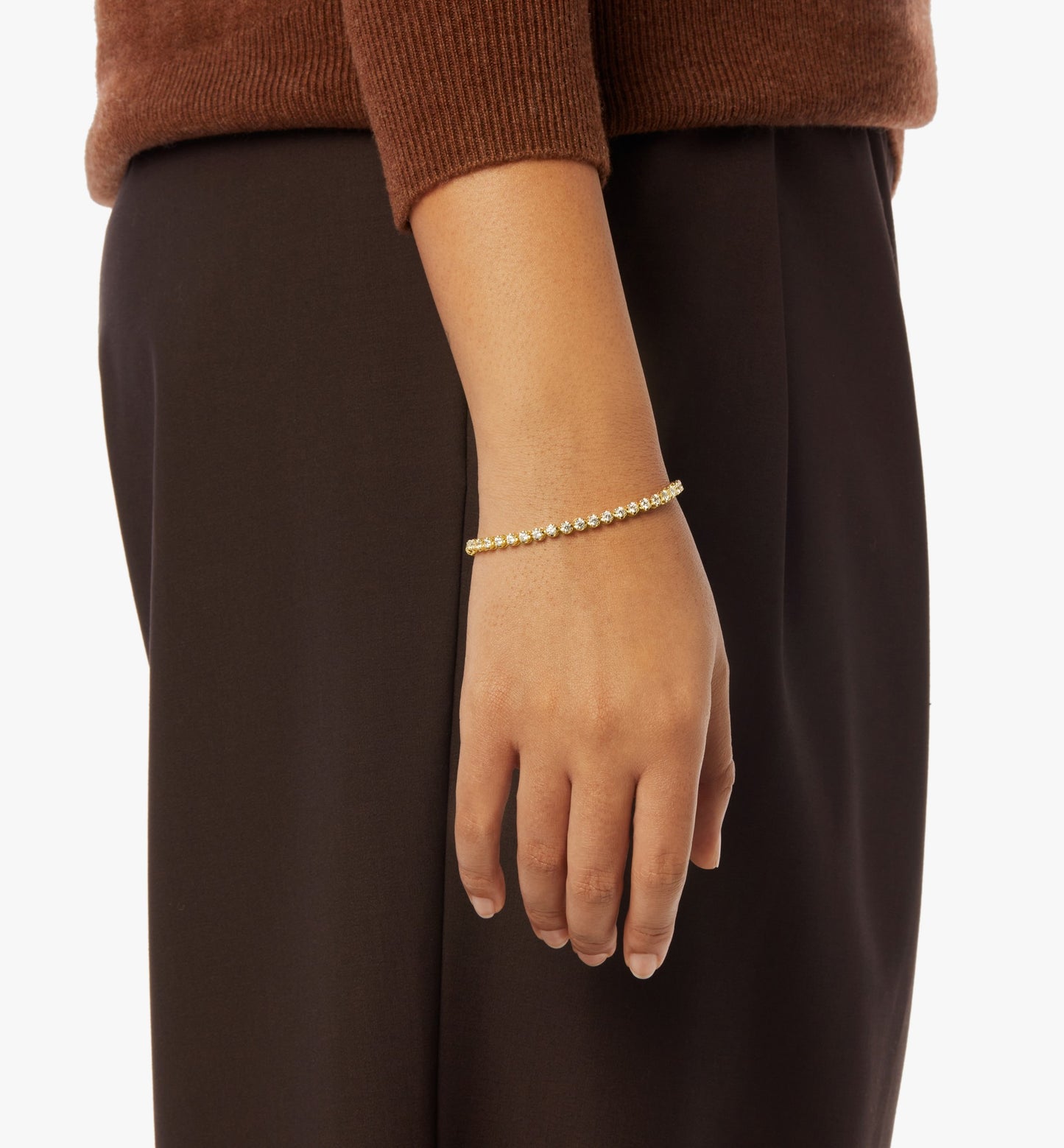 A close-up shows a person in brown pants and a brown sweater, their arm relaxed, highlighting the Shinola 4 Carat 7" Diamond Tennis Bracelet in yellow gold on their wrist.