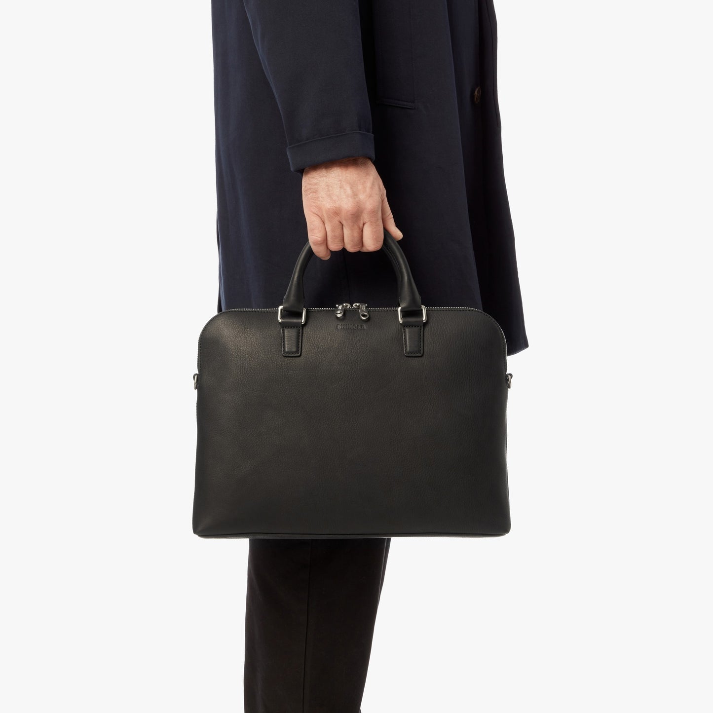 A person in a dark coat and pants holds the Shinola Portfolio Briefcase - Black by its handles, standing against a plain white background.