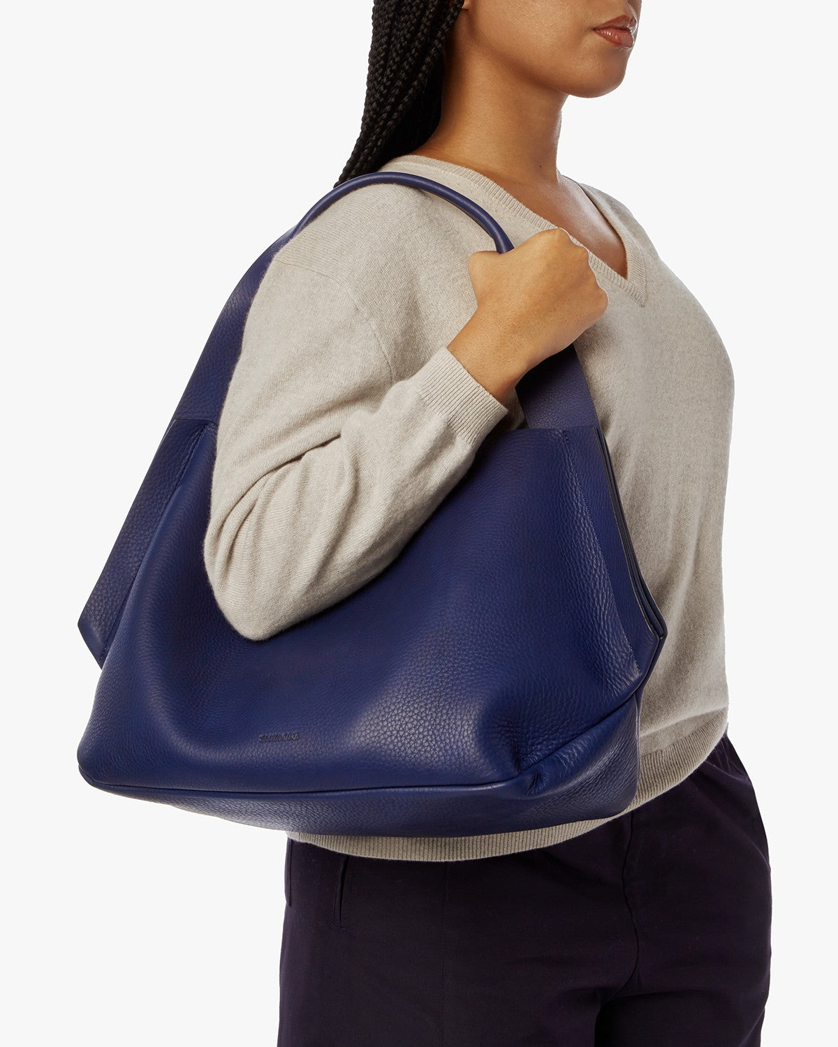 A person in a light beige sweater and dark pants carries the Shinola Runwell Shoulder Bag - Lake by its handles, resting it on their shoulder against a white background.