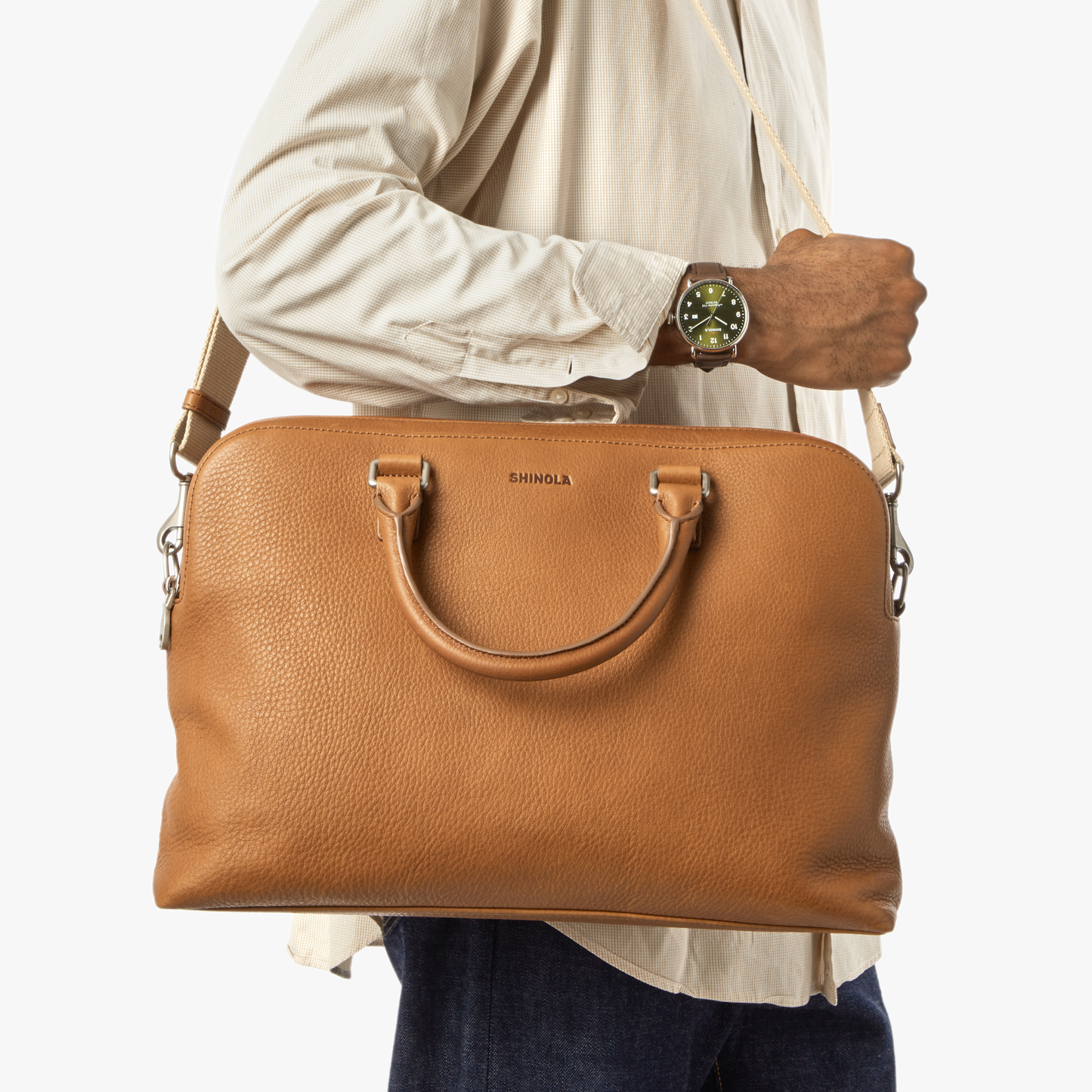 A person in a cream shirt and dark jeans carries a Shinola Double Zip Briefcase - Tan over their shoulder, holding the strap. They wear a green wristwatch and stand against a white background.