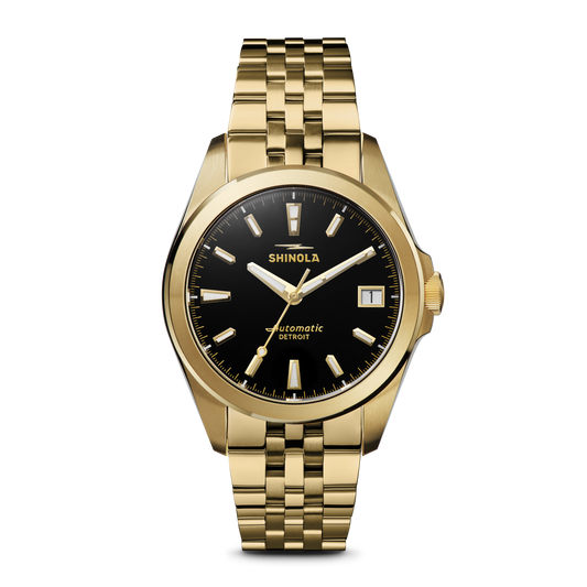 The Shinola Circadian Monster Automatic Watch in gold features a black dial, gold hour markers and hands, a date window at 3 o’clock, and a bold gold bracelet. The watch face reads "Shinola Automatic Detroit.