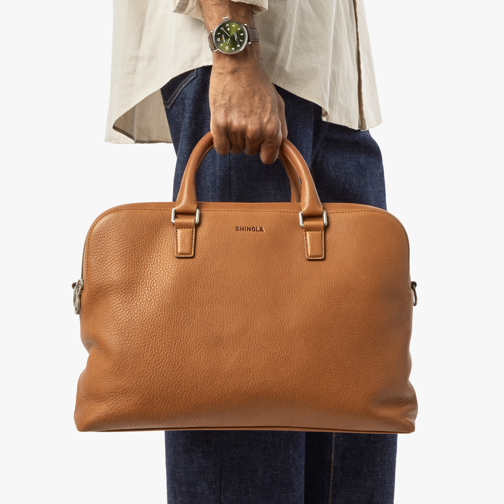 A person in a cream shirt, blue jeans, and a watch holds the Shinola Double Zip Briefcase - Tan by its handles, highlighting its extra compartment, set against a plain white background.