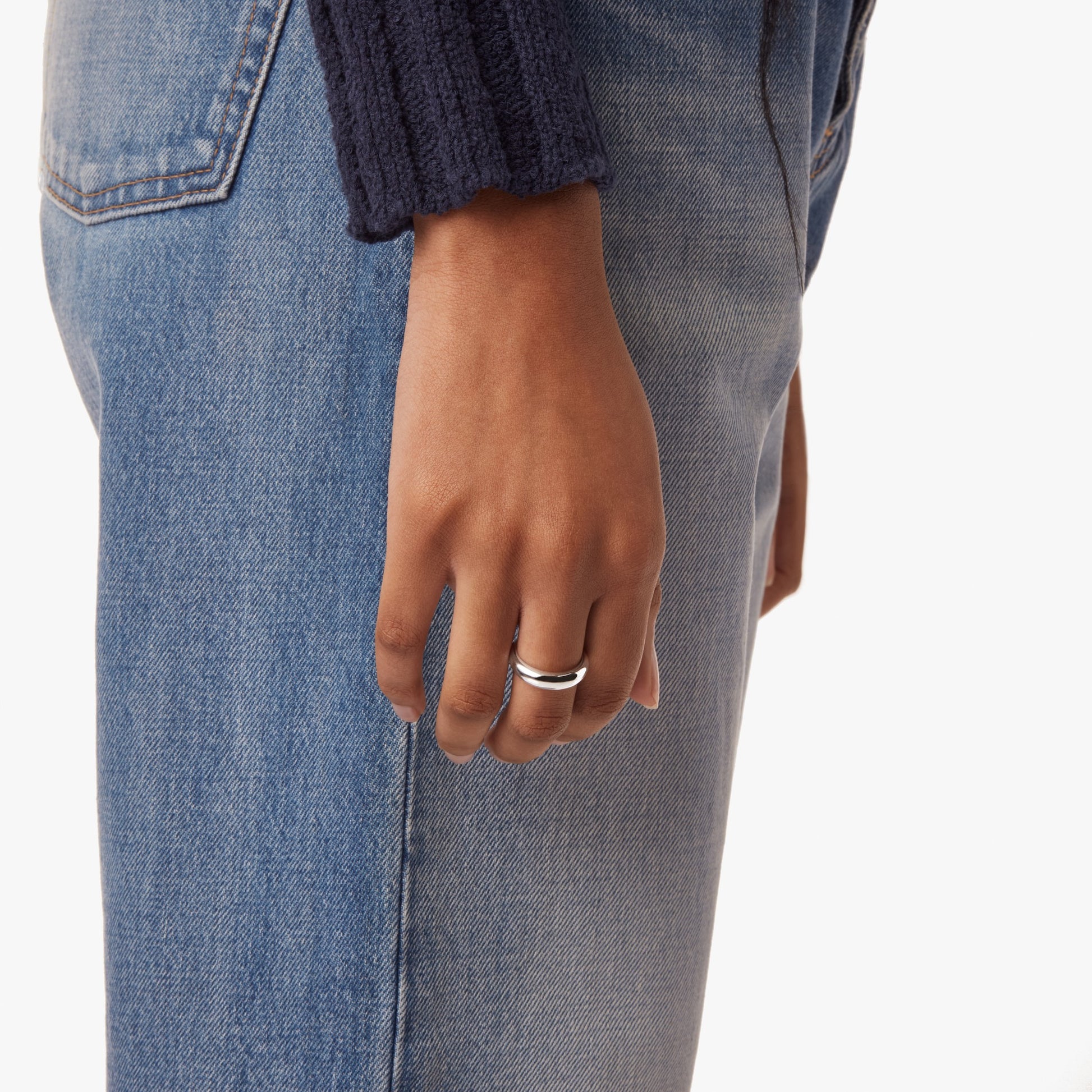 A person's hand, wearing the Shinola Signature Link Domed Ring - Sterling Silver on the middle finger, rests by their side. They wear blue jeans and a dark ribbed sweater, standing against a plain white background.
