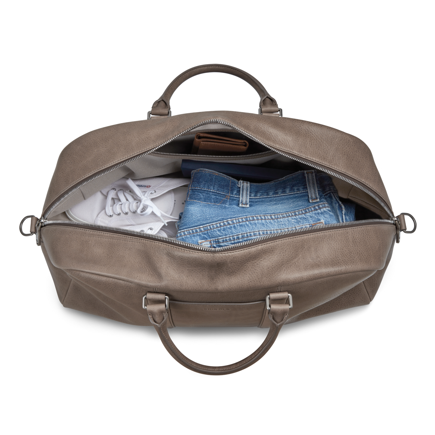 The Shinola Runwell Duffle Bag - Stone, crafted from natural grain leather, unzips to reveal a pair of white sneakers and folded blue denim jeans—combining style and function for the perfect carry-on.