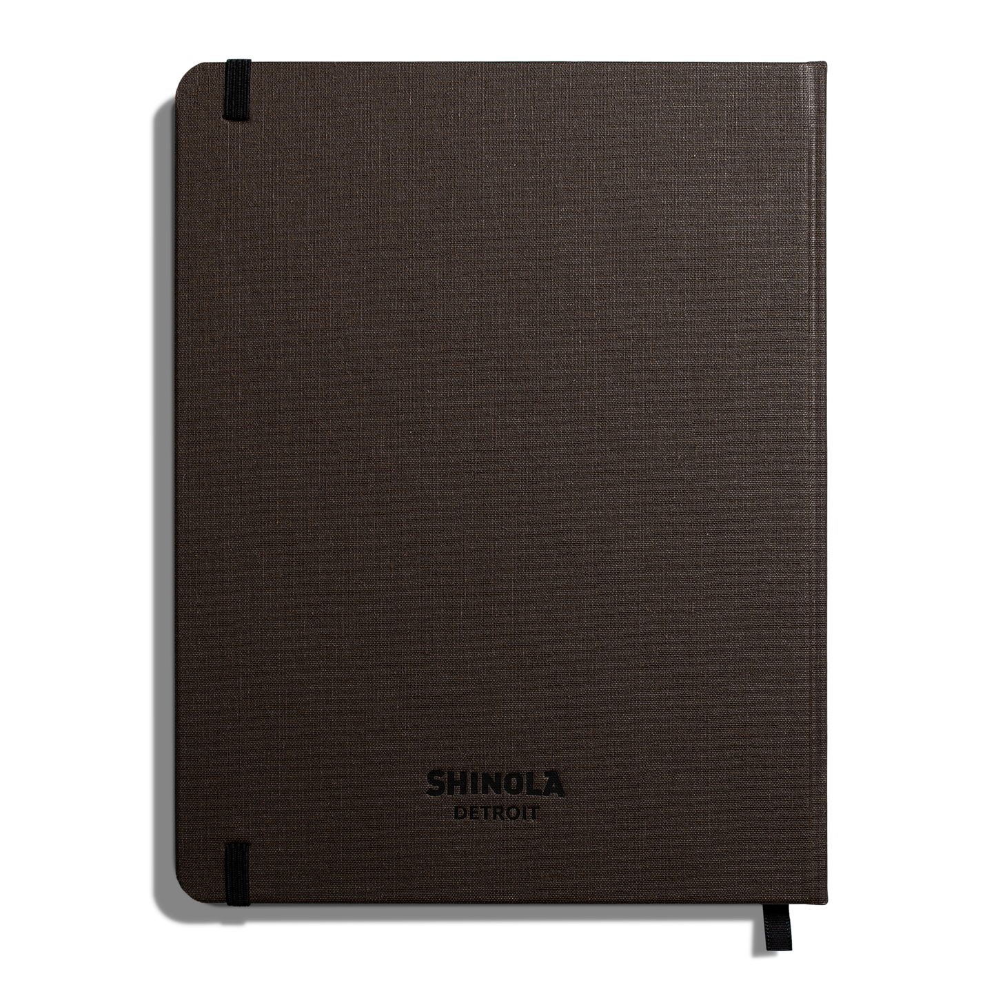 The Large Hard Ruled Journal - Espresso by Shinola is a closed, dark brown hardcover notebook with embossed branding, Smyth-sewn binding, and black elastic bands on the top left and bottom right corners.