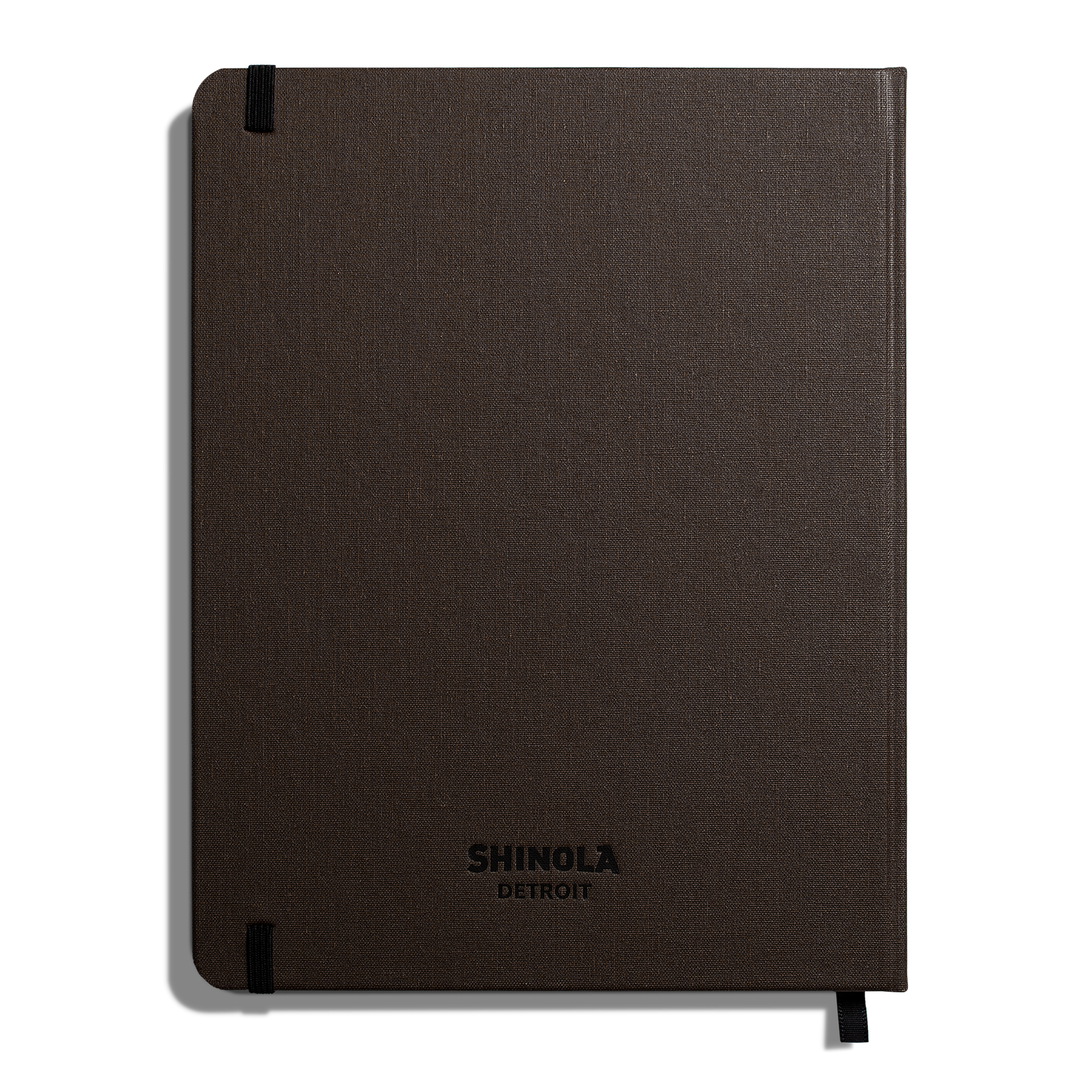 The Large Hard Ruled Journal - Espresso by Shinola is a closed, dark brown hardcover notebook with embossed branding, Smyth-sewn binding, and black elastic bands on the top left and bottom right corners.