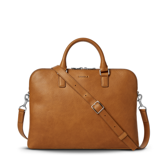 The Portfolio Briefcase - Tan by Shinola features top handles, a detachable shoulder strap, silver hardware, and an embossed logo on tan European leather. This slim laptop briefcase is showcased against a plain white background.