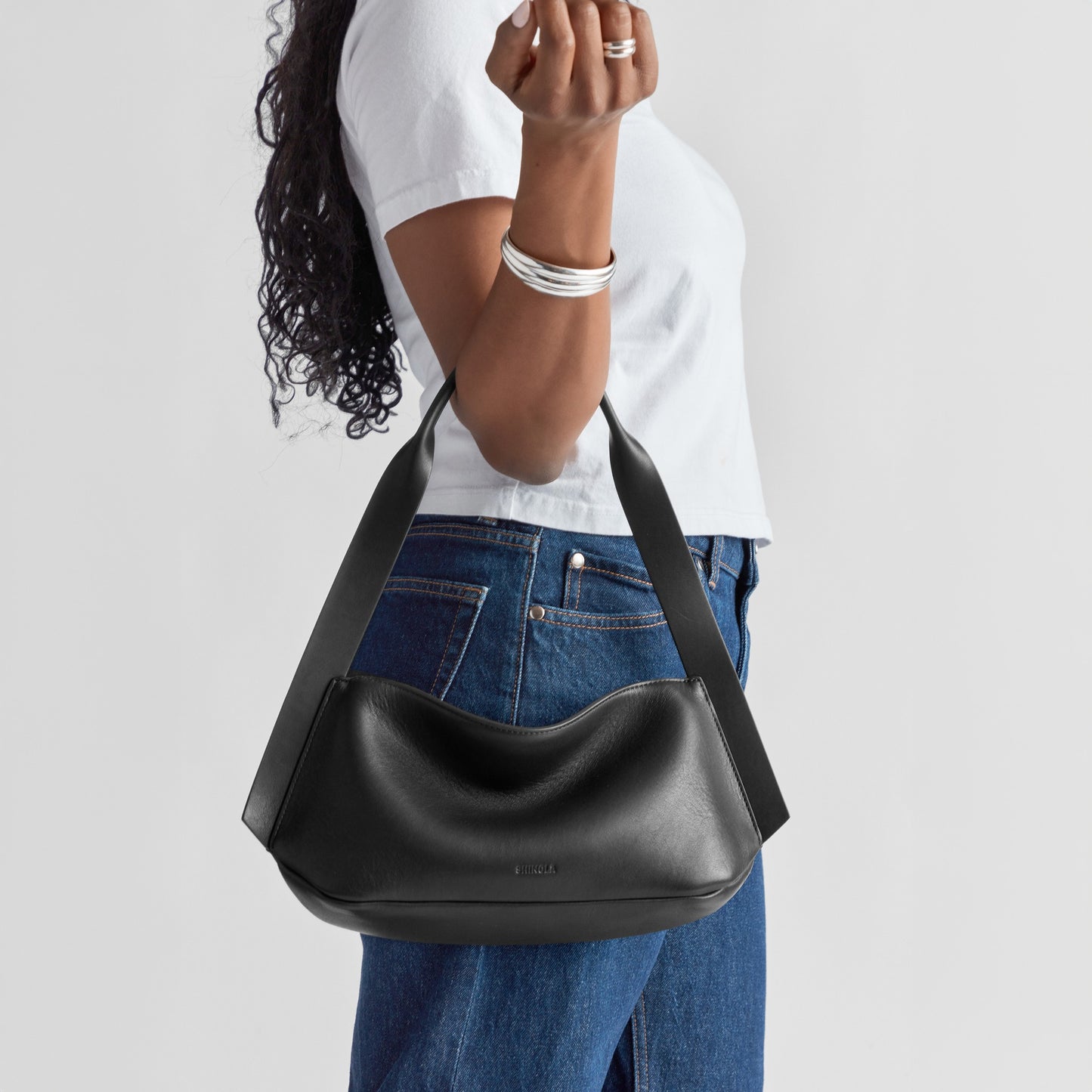 A person in a white t-shirt and blue jeans holds the Shinola Small Derby Shoulder Bag - Black, featuring natural leather and a wide strap. Their face is hidden, accessorized with silver bangles and a ring—ideal for day to night use.