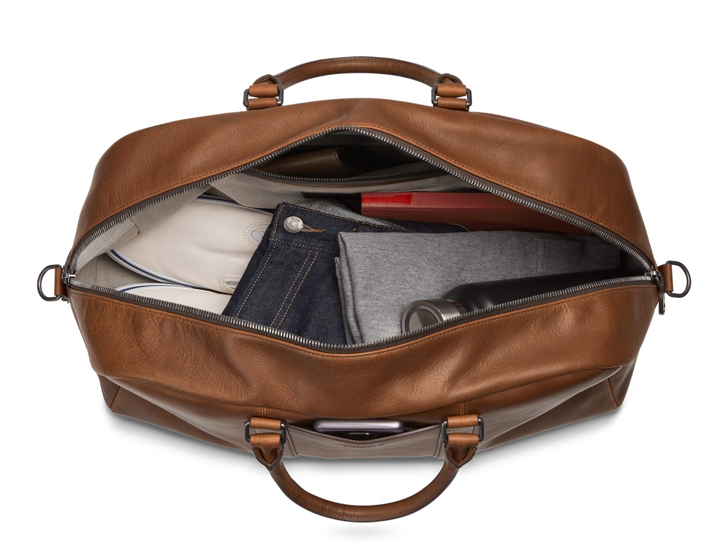 The Shinola Runwell Duffle Bag - Medium Brown is unzipped to show neatly packed clothes, a water bottle, and a red rectangular item inside. The bag is viewed from above against a plain white background.