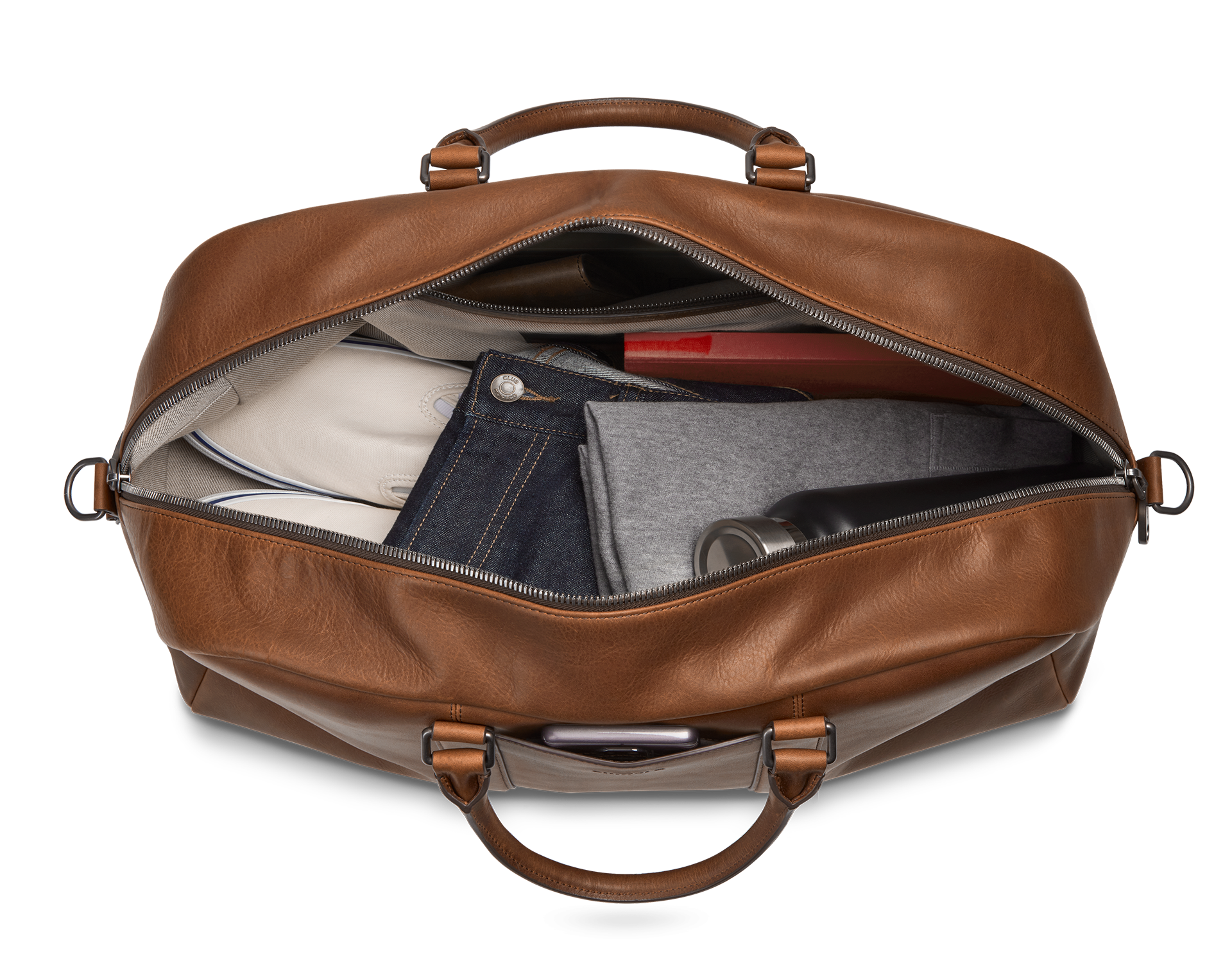 The Shinola Runwell Duffle Bag - Medium Brown is unzipped to show neatly packed clothes, a water bottle, and a red rectangular item inside. The bag is viewed from above against a plain white background.