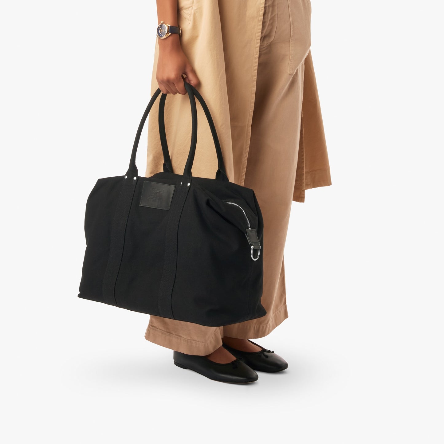 A person in beige pants and black flats holds the Shinola Runabout Holdall Canvas Bag - Black by its handles. The image shows only their lower half against a plain white background.
