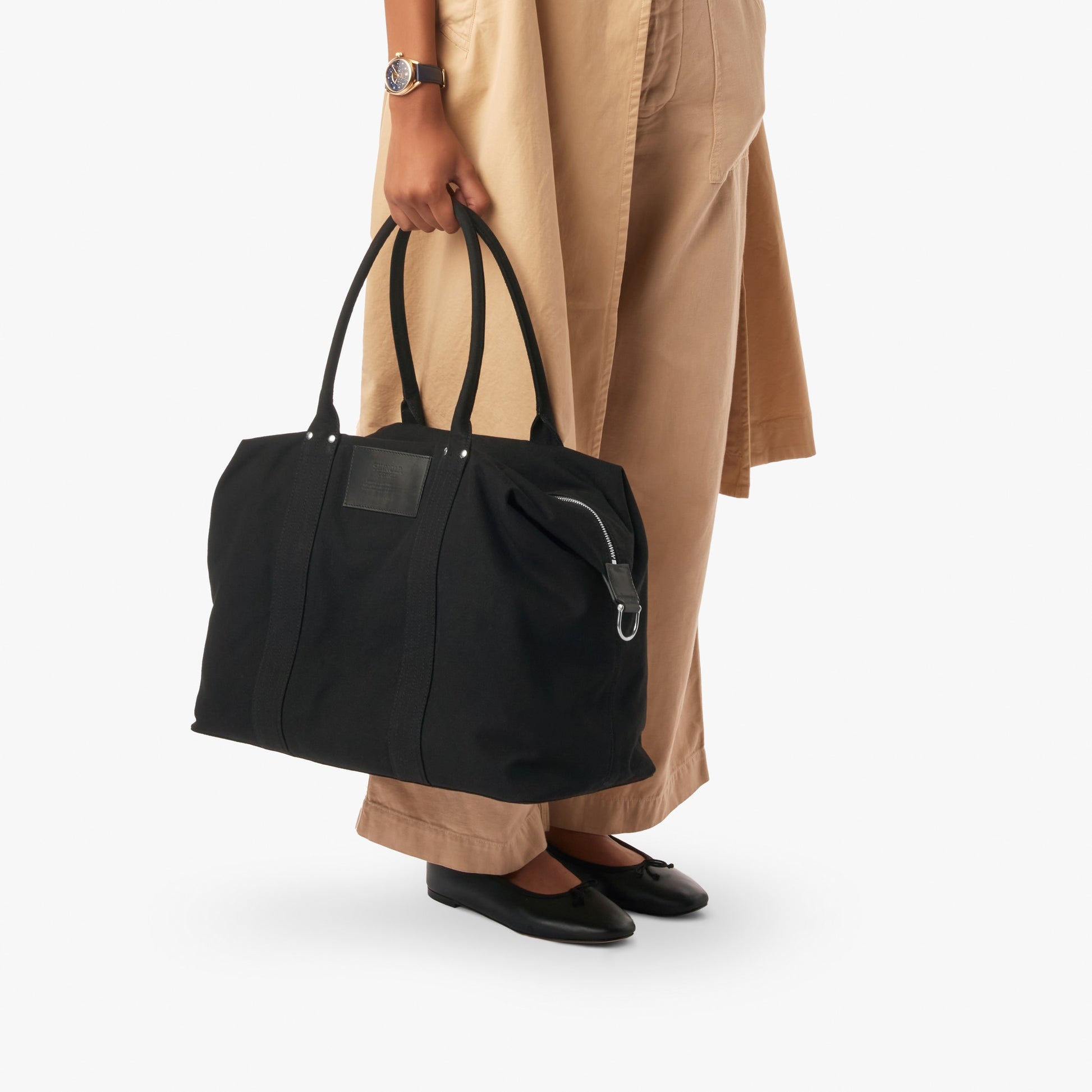 A person in beige pants and black flats holds the Shinola Runabout Holdall Canvas Bag - Black by its handles. The image shows only their lower half against a plain white background.