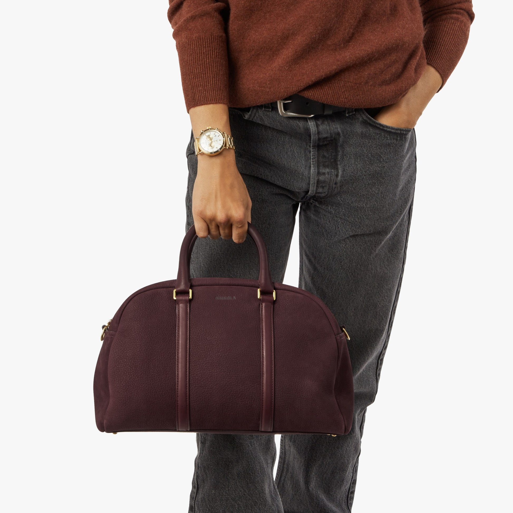 A person in a rust sweater and dark jeans holds the Shinola Runwell Satchel - Merlot by its handles, with their watch and belt visible against a plain white background.
