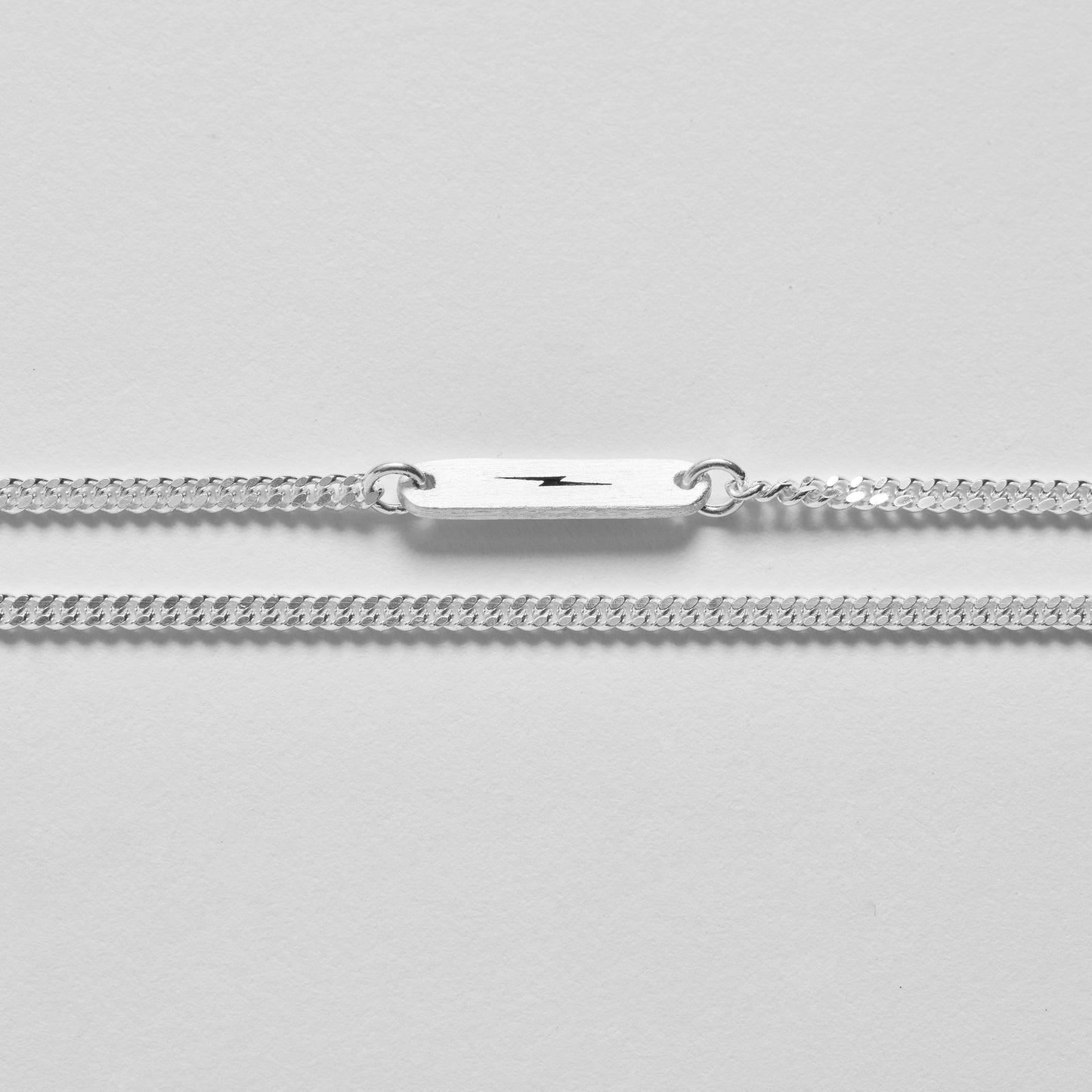 A sterling silver chain bracelet with a black lightning bolt plate is shown on a light gray background. Style it with the Shinola Simple Curb Chain Necklace in sterling silver for a timeless look.
