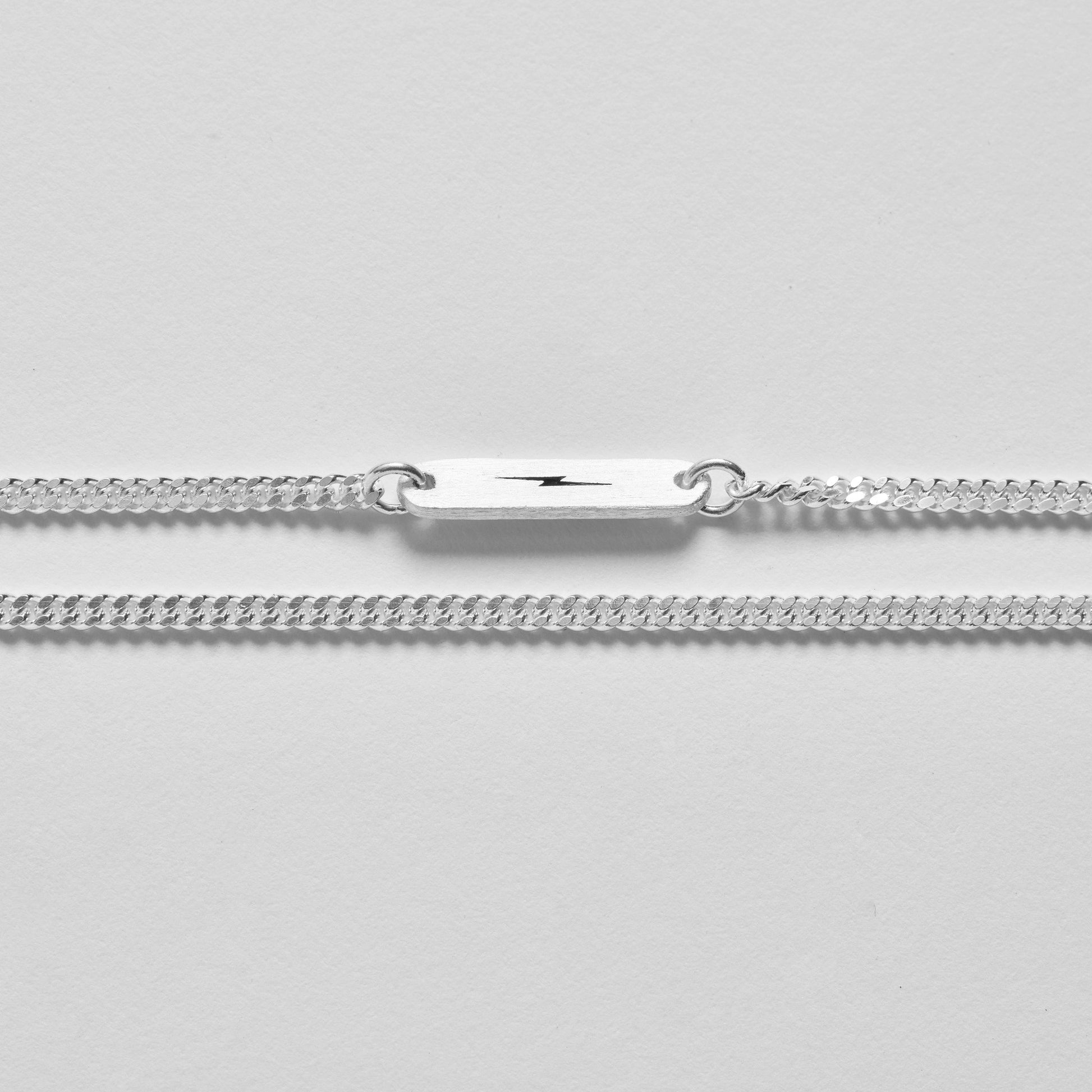 A sterling silver chain bracelet with a black lightning bolt plate is shown on a light gray background. Style it with the Shinola Simple Curb Chain Necklace in sterling silver for a timeless look.