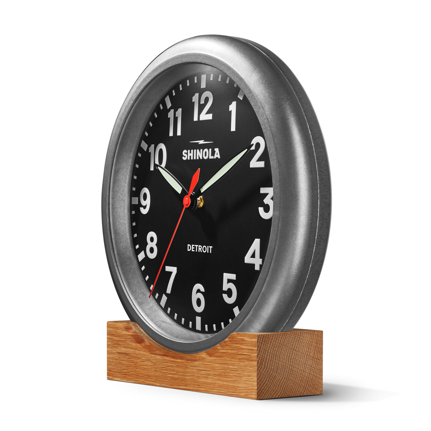 The Shinola 14" Runwell Clock in Tumbled Zinc with a black face, white numbers, and white hands features a round silver frame and sits on a wooden block base against a plain white background, illustrating classic industrial style.