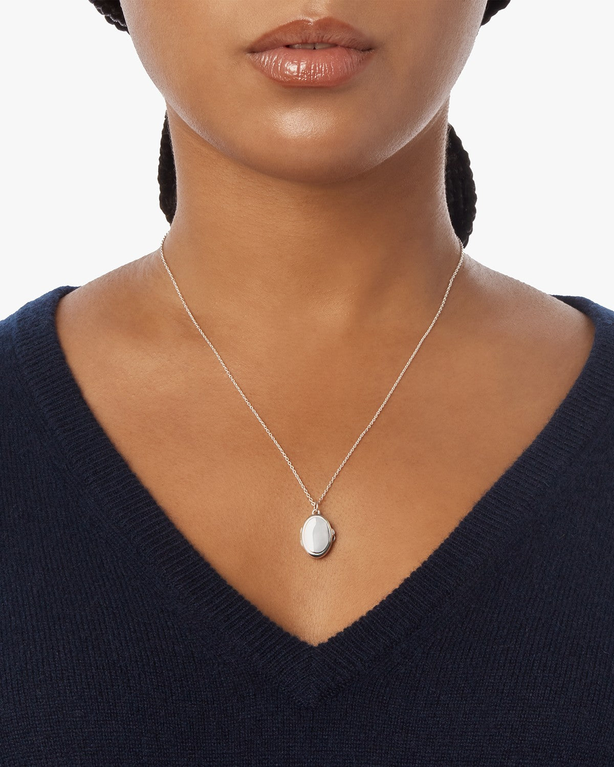 A woman wearing a navy V-neck sweater showcases the Shinola Heirloom Petite Locket Necklace in sterling silver, its oval pendant visible, photographed from shoulders to chin against a plain white background.