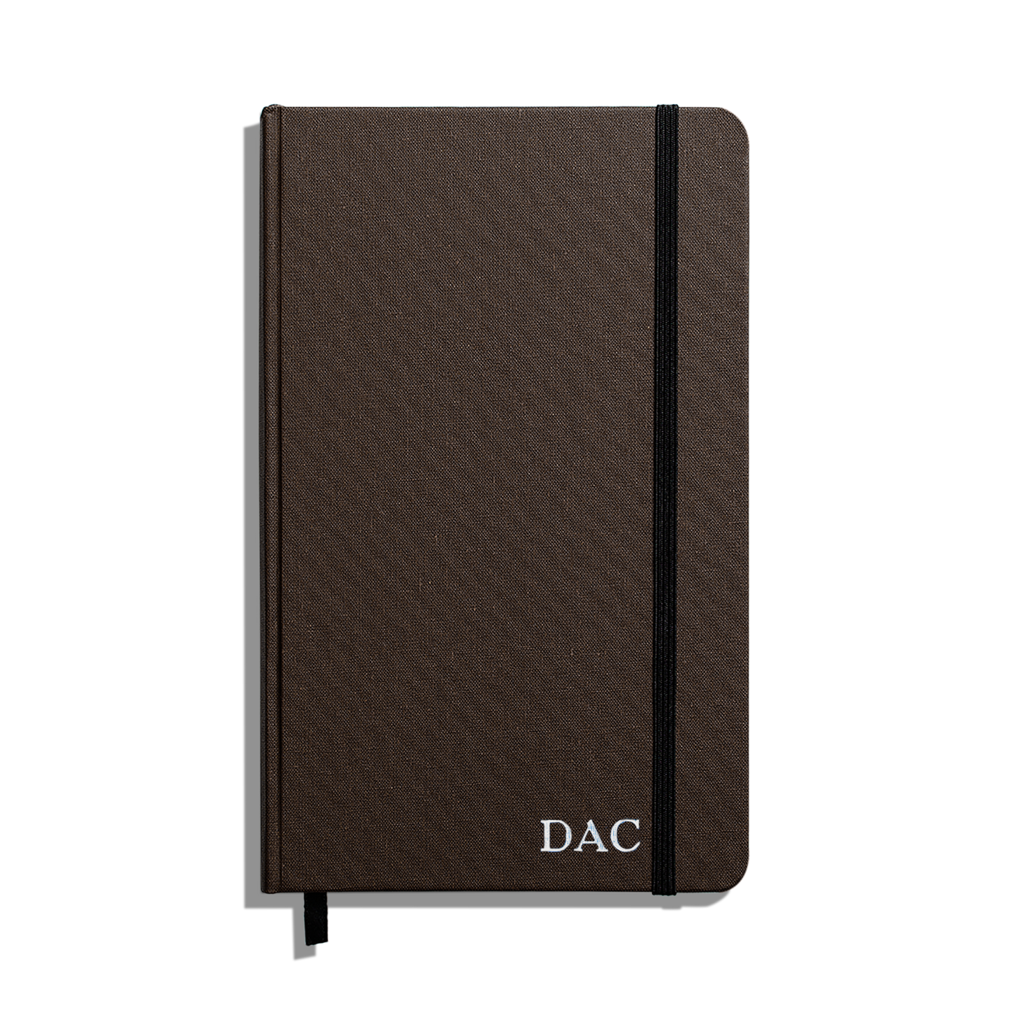 The Shinola Medium Hard Ruled Journal (5.25" x 8.25") is a closed brown notebook with a black elastic band, ribbon bookmark, and white "DAC" initials on the cover, featuring durable Smyth-sewn binding and acid-free pages.