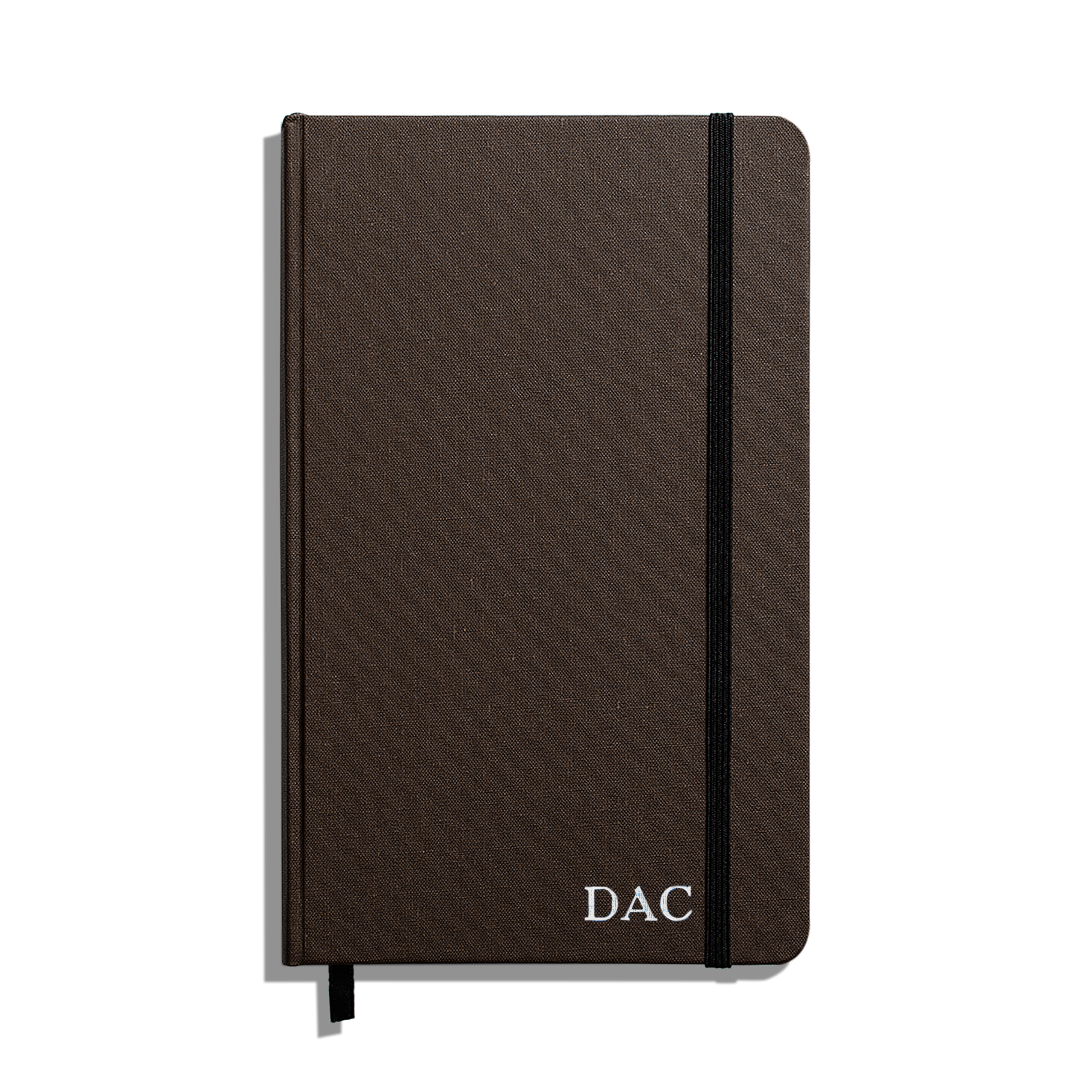 The Shinola Medium Hard Ruled Journal (5.25" x 8.25") is a closed brown notebook with a black elastic band, ribbon bookmark, and white "DAC" initials on the cover, featuring durable Smyth-sewn binding and acid-free pages.