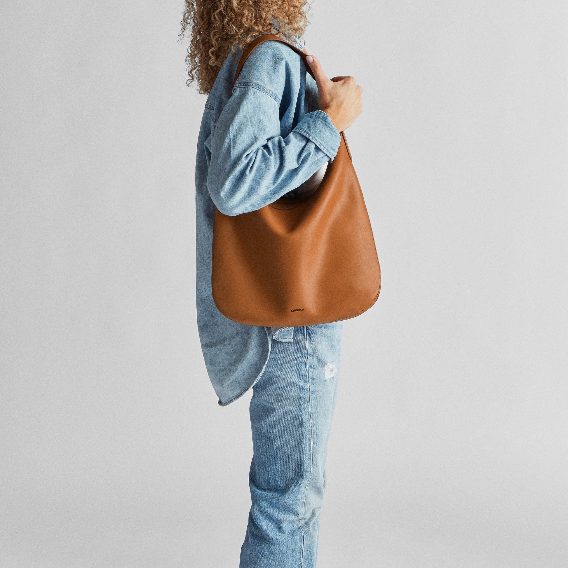 A person with curly hair, in a loose denim shirt and jeans, stands sideways holding the Shinola Bixby Shoulder Bag - Tan, featuring natural leather and accordion-style construction, against a plain light background.