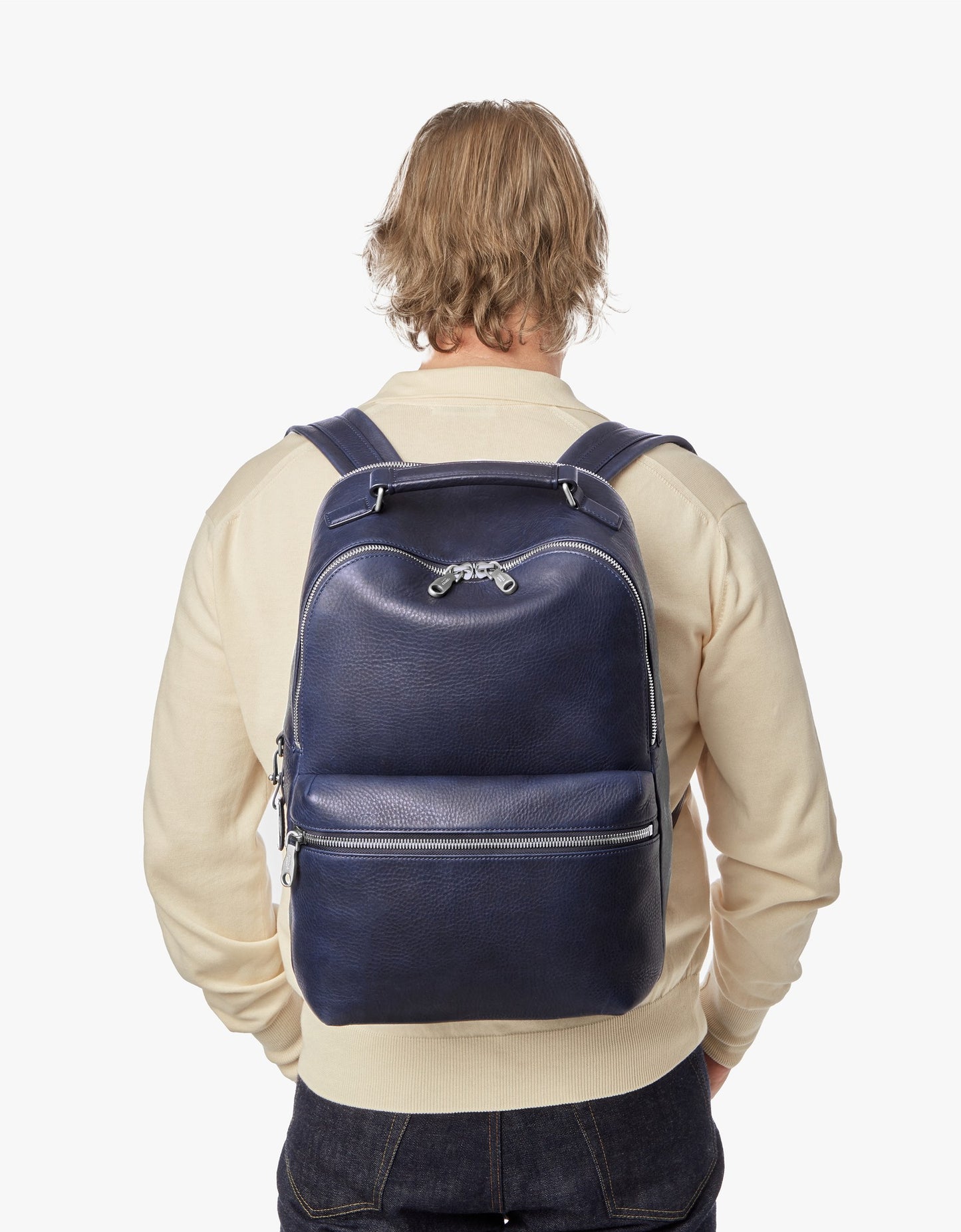 A person with blond hair, wearing a cream sweater and dark jeans, is seen from behind carrying the Shinola Portfolio Briefcase - Stone against a plain white background.